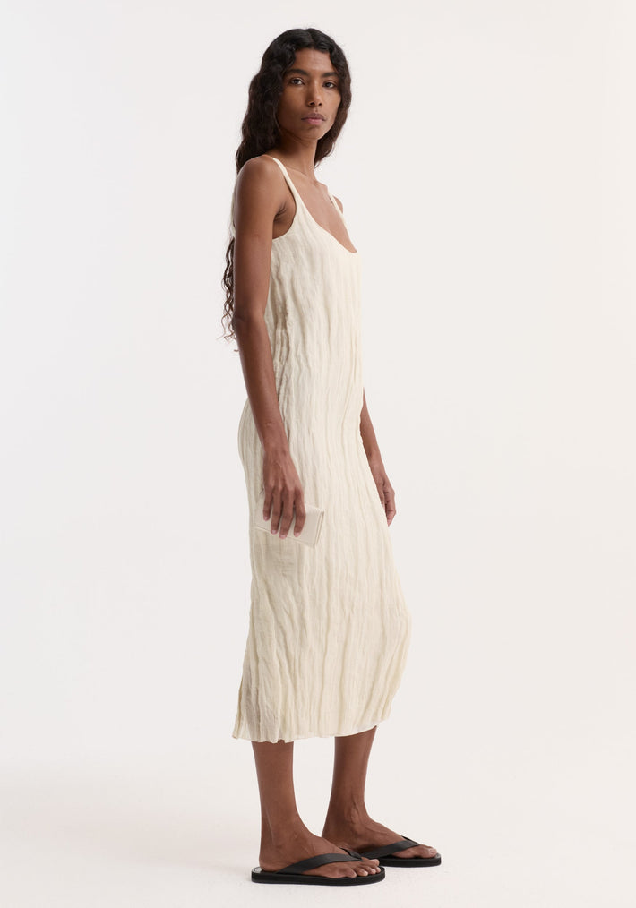 Woman in a Textured square neck dress | cream, sleeveless with a scoop neck, holding a small item, showcasing the dress's crinkled texture and straight silhouette.