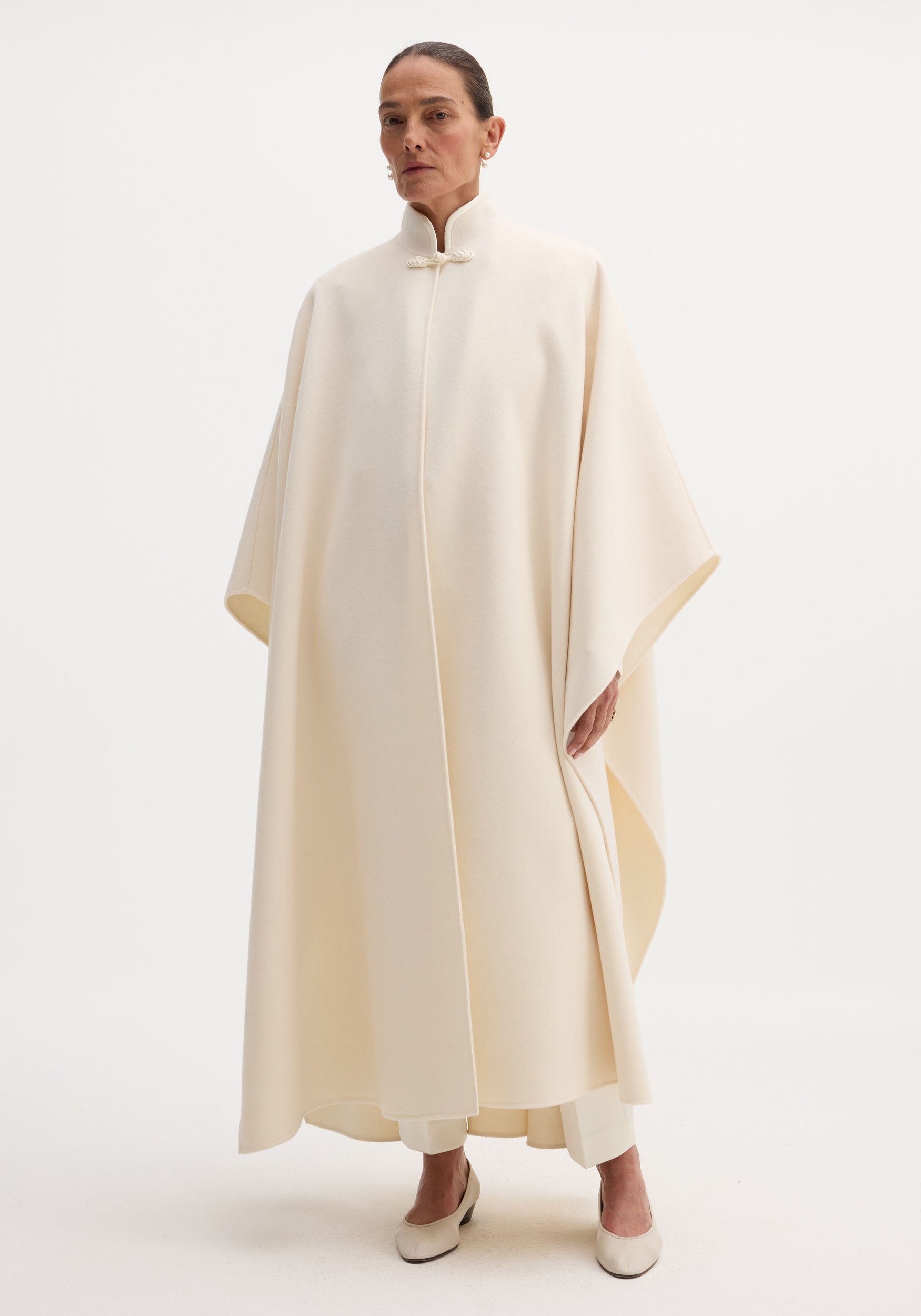 A model wearing a minimalist off white Pankou closure poncho with a high collar and elegant draping, styled with matching off white pants and shoes.