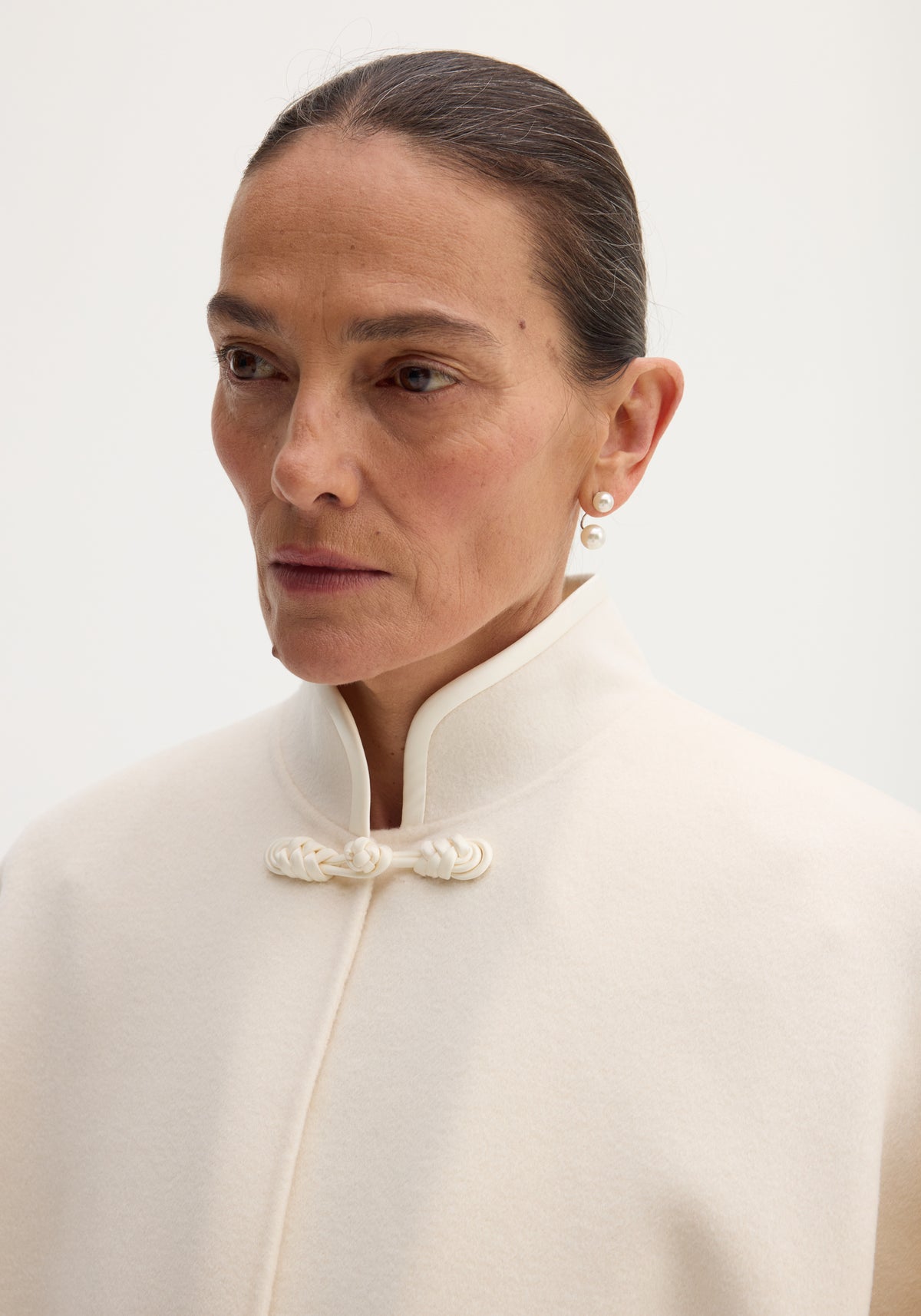 Close-up of a woman wearing a high-collar off white poncho with a pankou closure detail, paired with elegant pearl drop earrings, showcasing the minimalist style of the Pankou closure poncho | off white.