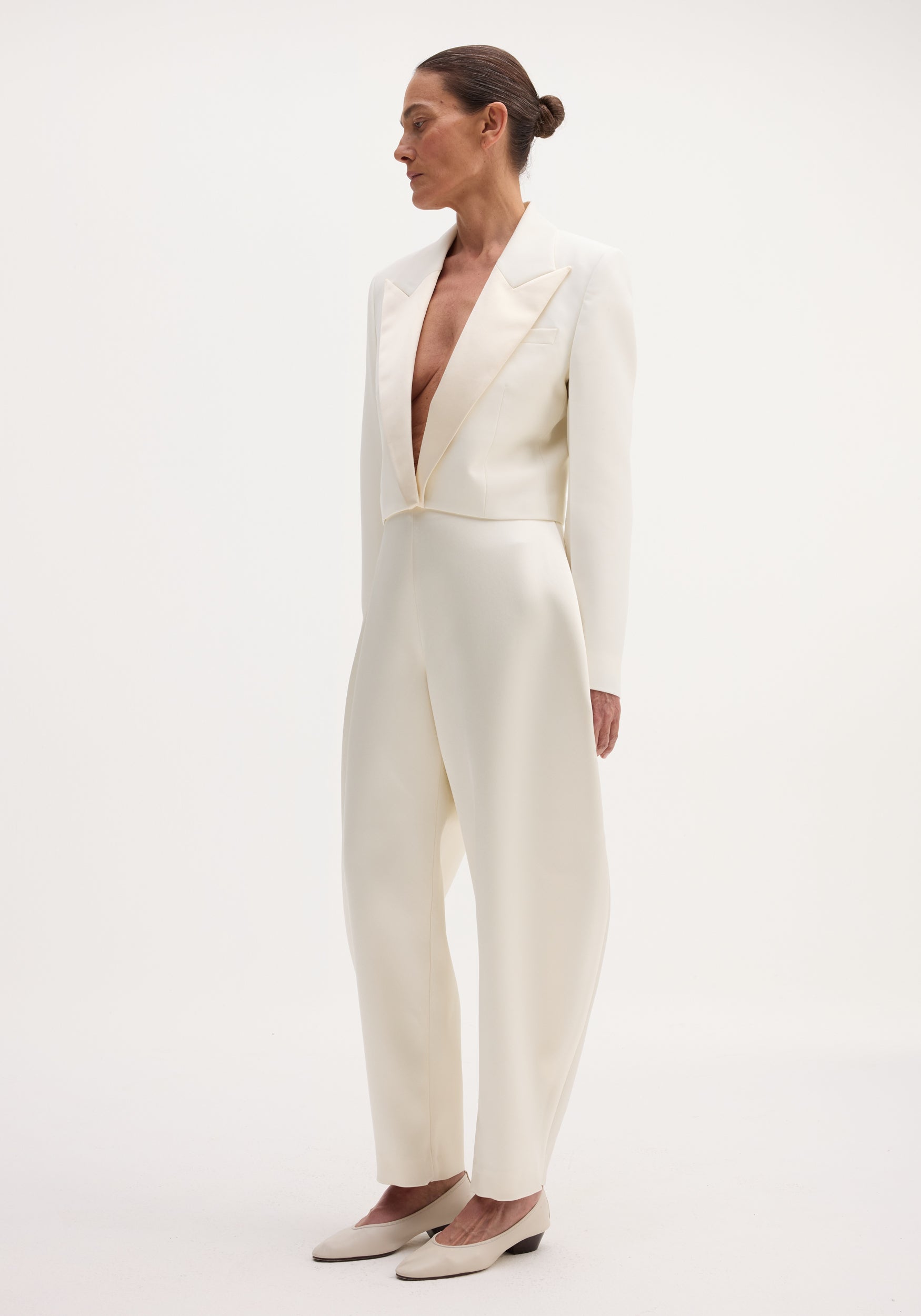 Woman wearing a tailored Cropped blazer | off white with deep V-neck and matching high-waisted trousers, styled with low-heeled shoes against a plain background.