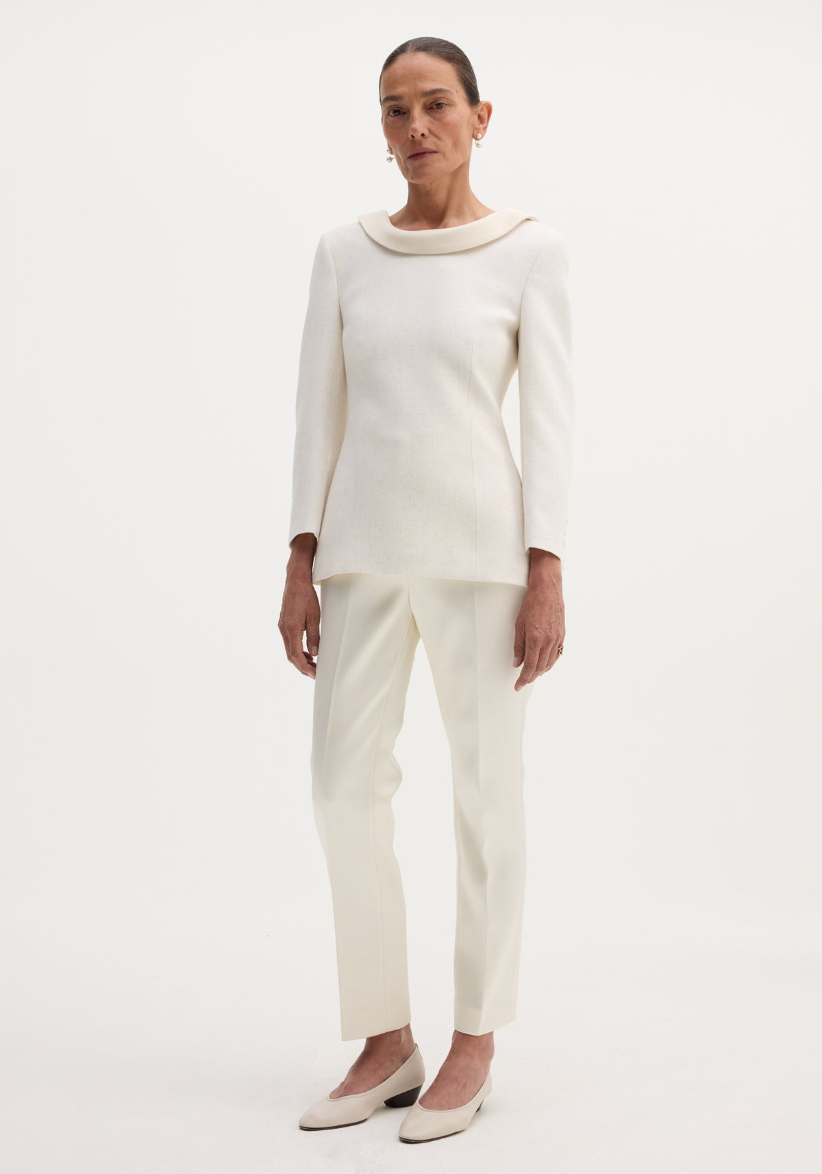 Woman wearing ivory cigarette leg satin trousers | ivory paired with a long-sleeve light ivory top and matching low-heeled shoes against a plain white background.