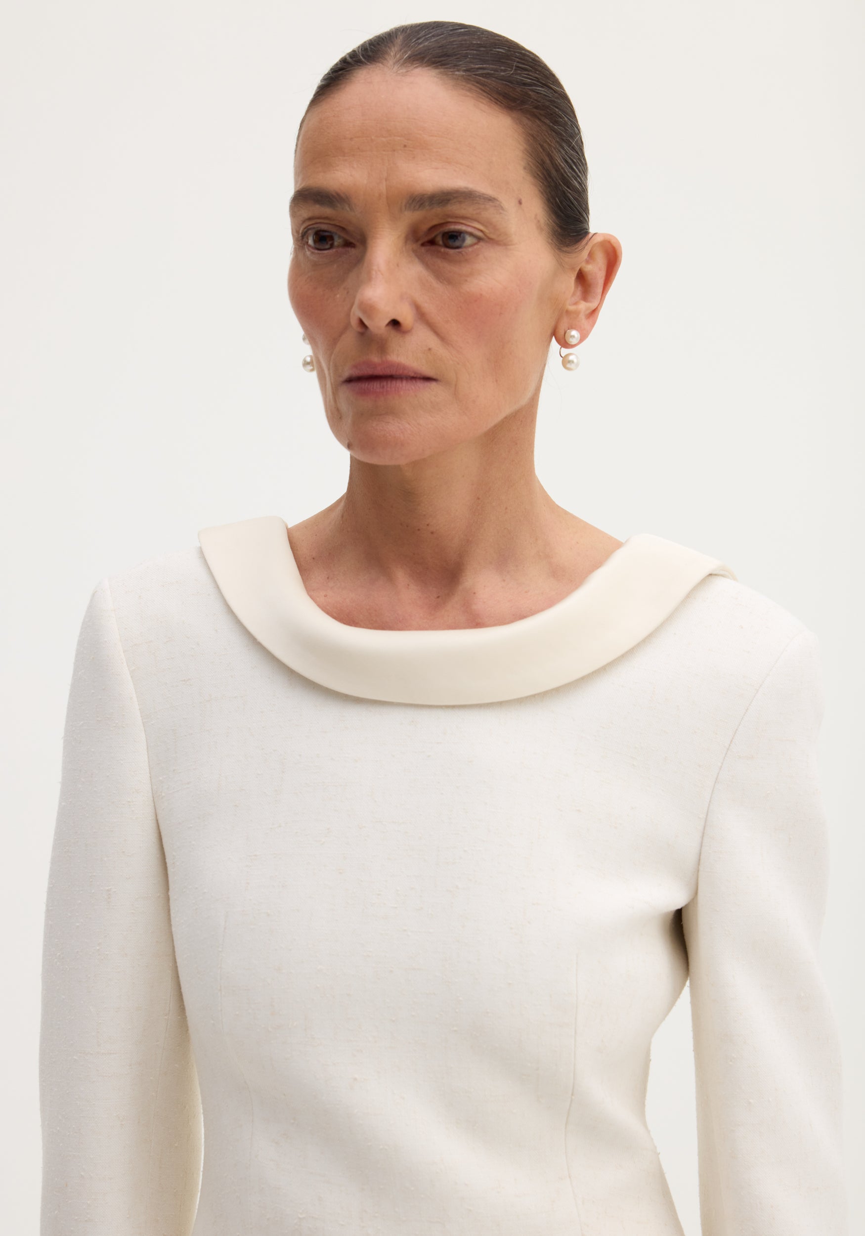 Woman wearing a minimalist Reversed blazer | winter white with a wide, turned collar and long sleeves, styled with pearl earrings against a plain light background.