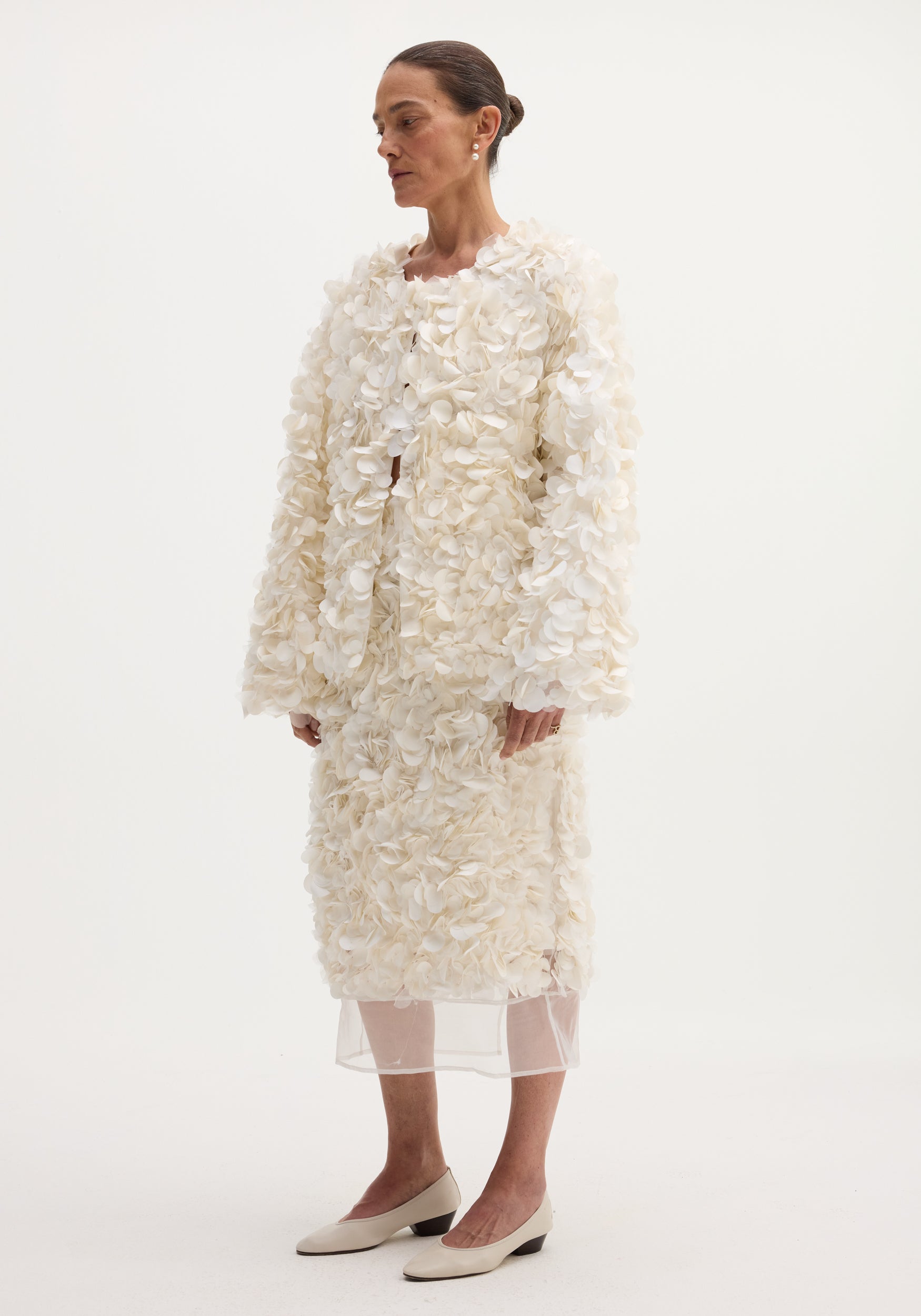 Woman wearing a handmade embellished jacket | ivory with 3D floral appliqués, paired with a matching skirt and beige low-heeled shoes against a plain background.