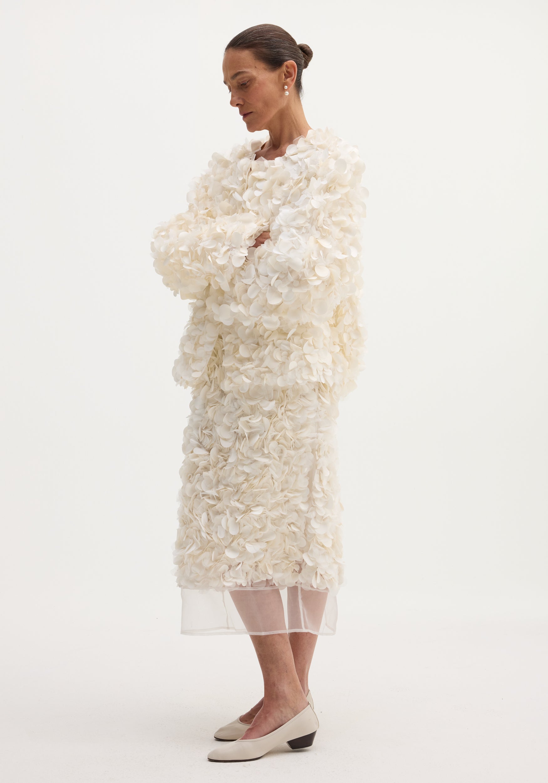 Woman wearing a long, textured jacket covered with layered fabric petals in ivory color, styled with a sheer underlayer and ivory low-heeled shoes, featuring the Handmade embellished jacket | ivory.