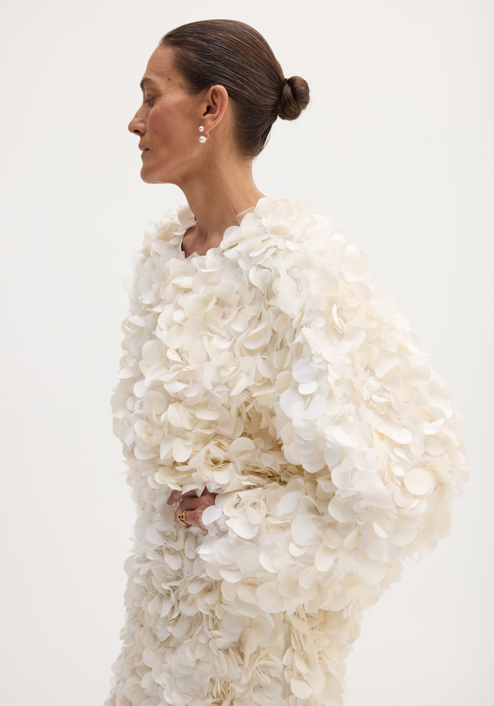 Side profile of a woman wearing a handmade embellished jacket | ivory adorned with layered floral appliqués, showcasing textured detail and voluminous sleeves against a plain background.