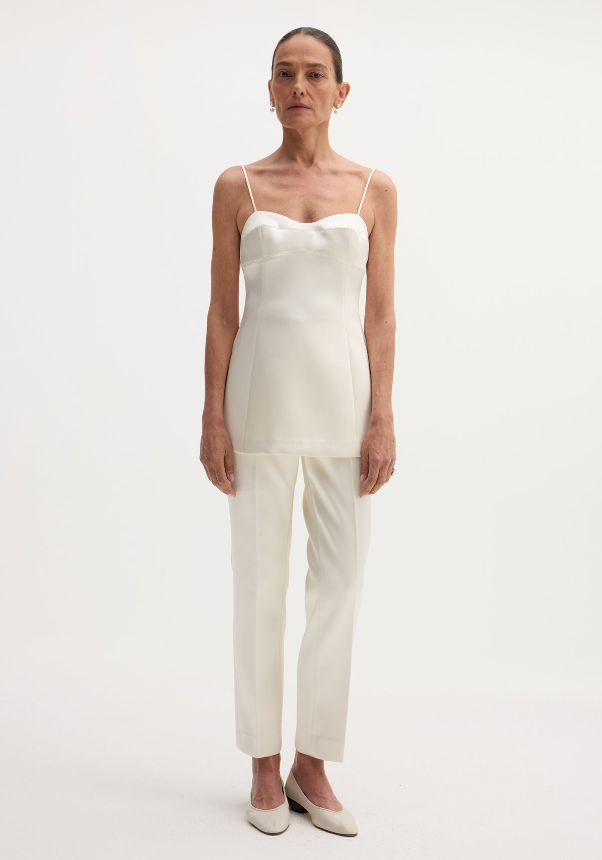 Woman wearing satin bustier top | ivory with thin straps, paired with matching ivory pants and low-heeled shoes, standing against a plain white background.