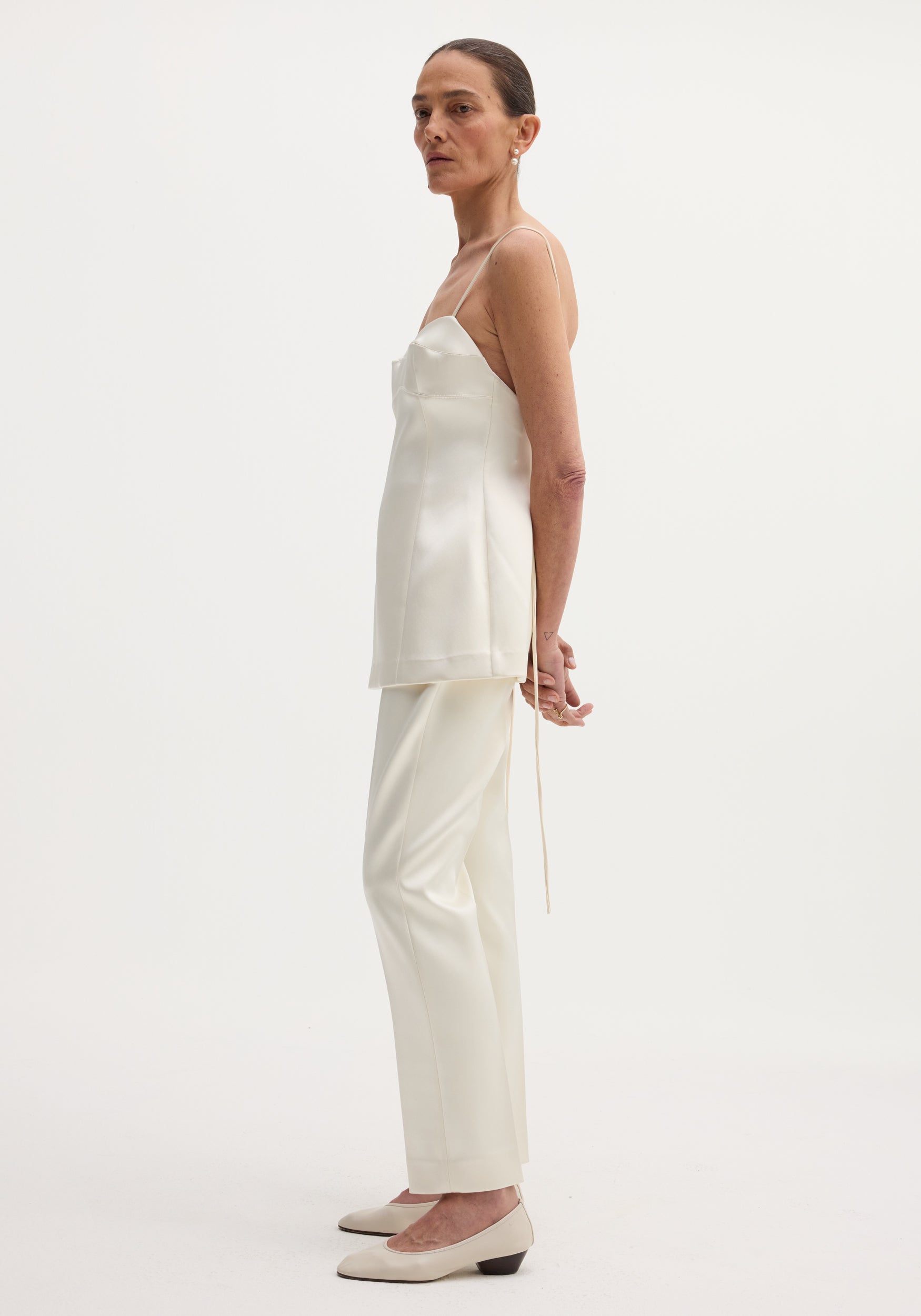 Woman wearing an elegant satin bustier top | ivory with thin straps, paired with matching ivory trousers and cream low-heeled shoes against a plain light background.