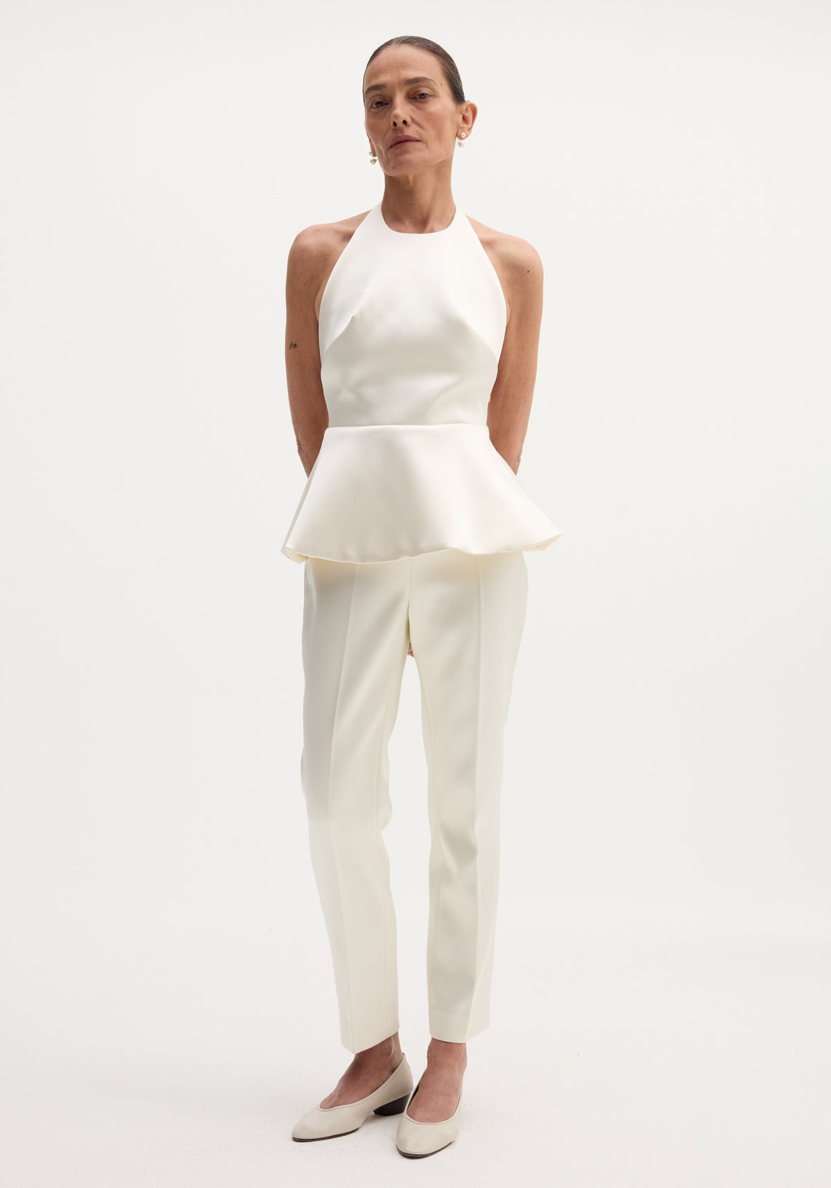 Woman wearing a sculptural satin halter top | ivory paired with matching tailored ivory pants and cream flats, standing against a plain white background.