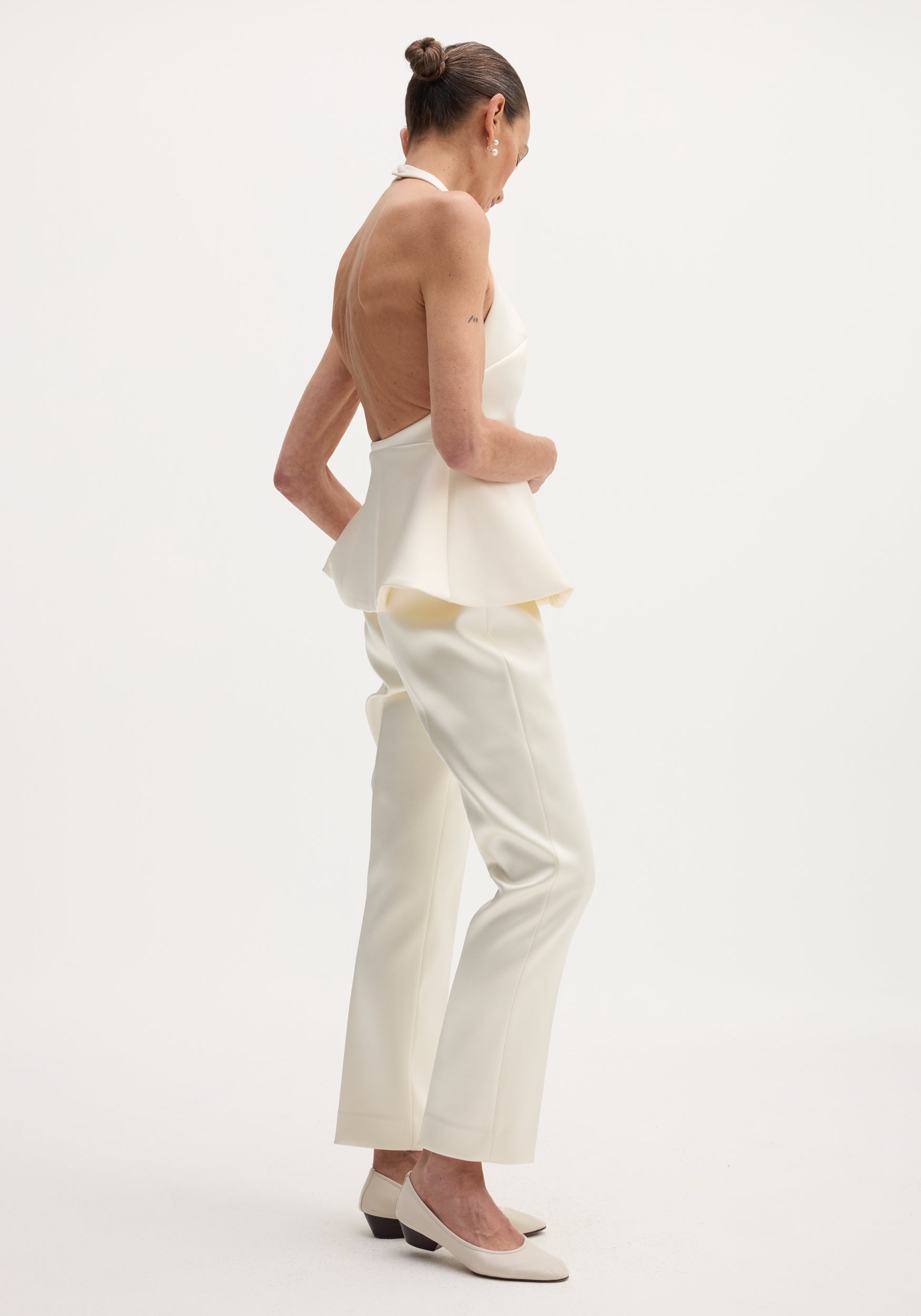 Woman wearing an ivory sculptural satin halter top with an open back and peplum detail, paired with matching ivory trousers and low-heeled shoes.