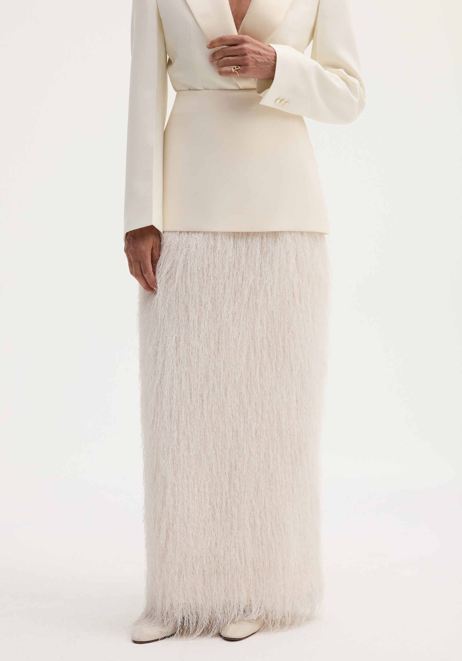 Person in ivory ensemble featuring Fringe skirt | ivory with tailored blazer; floor-length fringe adds movement. Cream shoes visible, hand adorned with a gold ring.