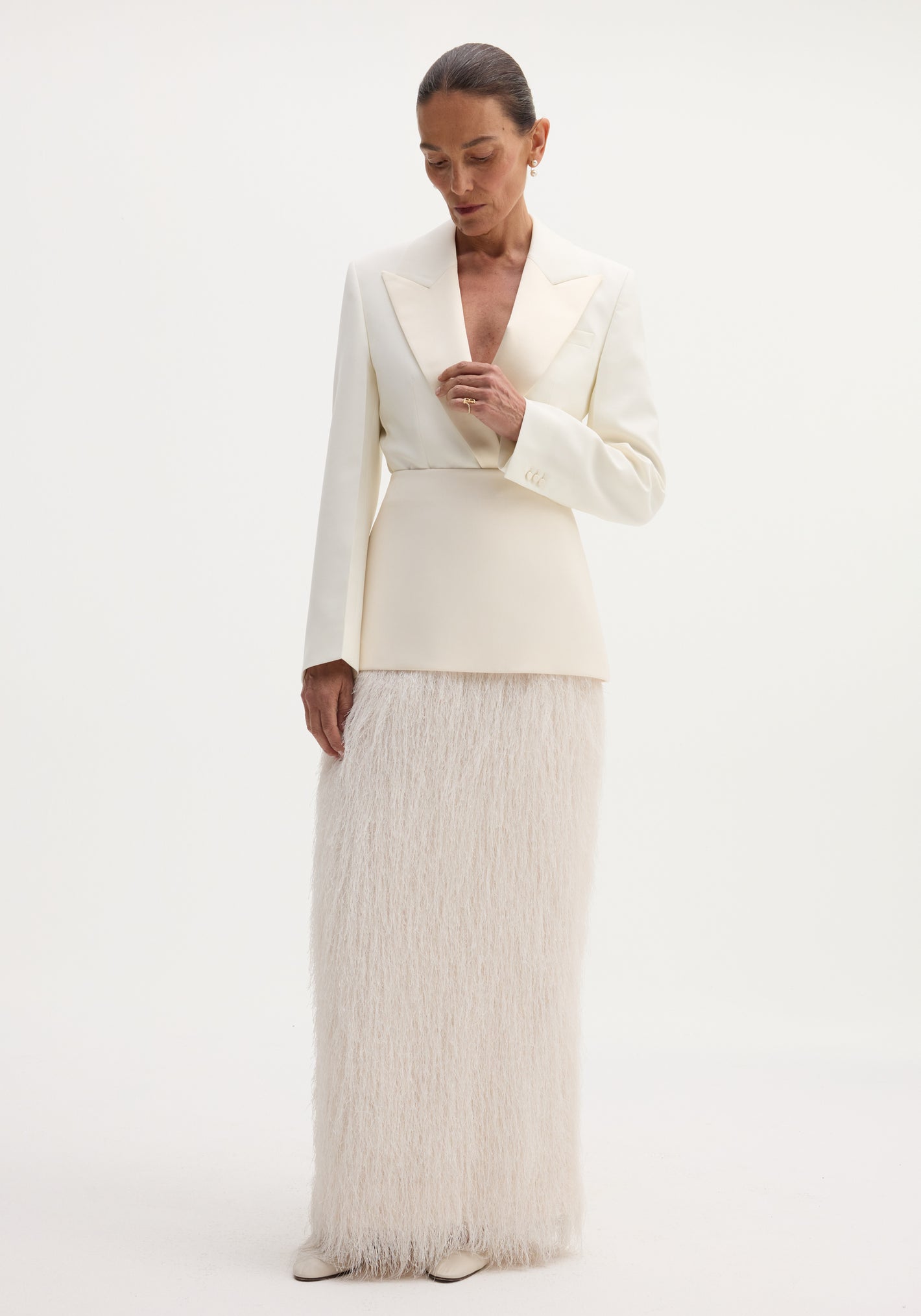 Model in an ivory Fringe skirt featuring a fitted wool upper and full-length fringe, paired with a tailored blazer, pearl earrings, and slicked-back hair.