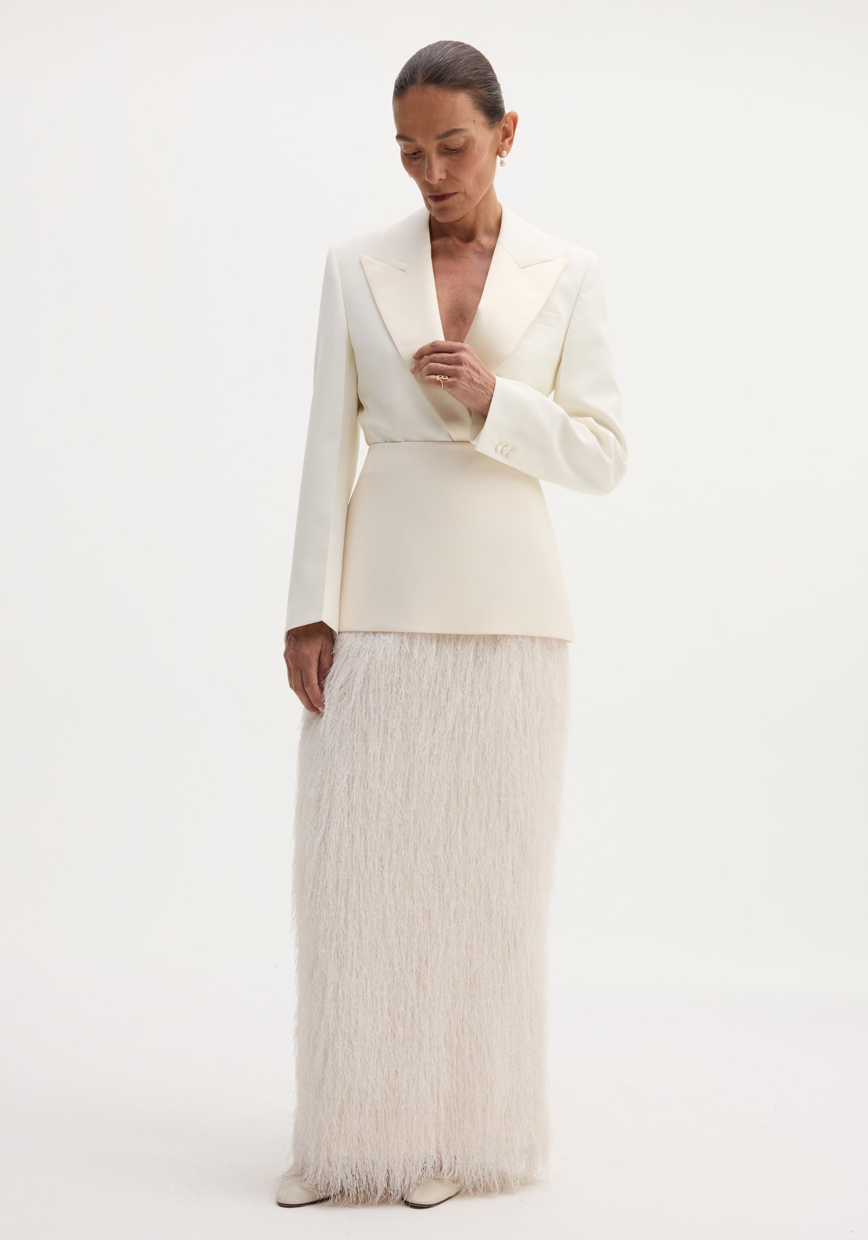Model in an ivory Fringe skirt featuring a fitted wool upper and full-length fringe, paired with a tailored blazer, pearl earrings, and slicked-back hair.
