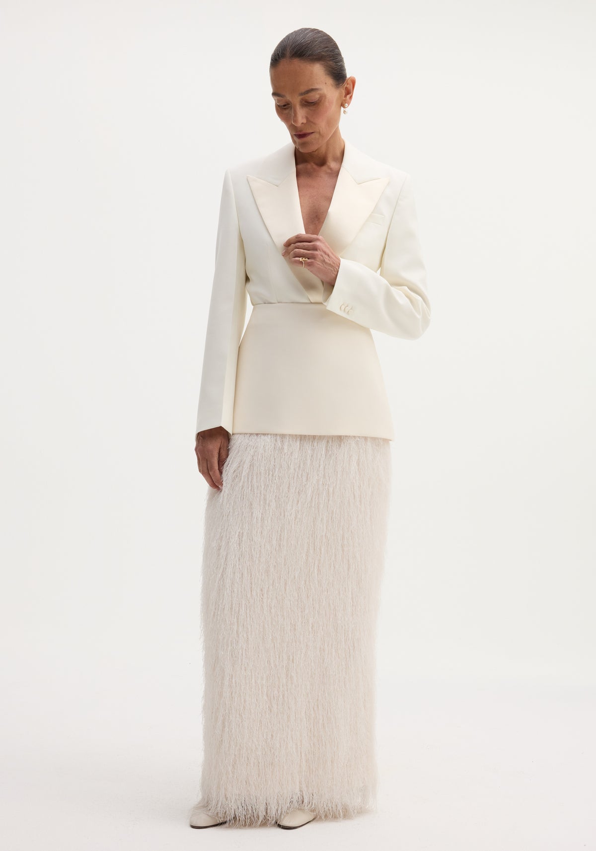 Model in an ivory Fringe skirt featuring a fitted wool upper and full-length fringe, paired with a tailored blazer, pearl earrings, and slicked-back hair.