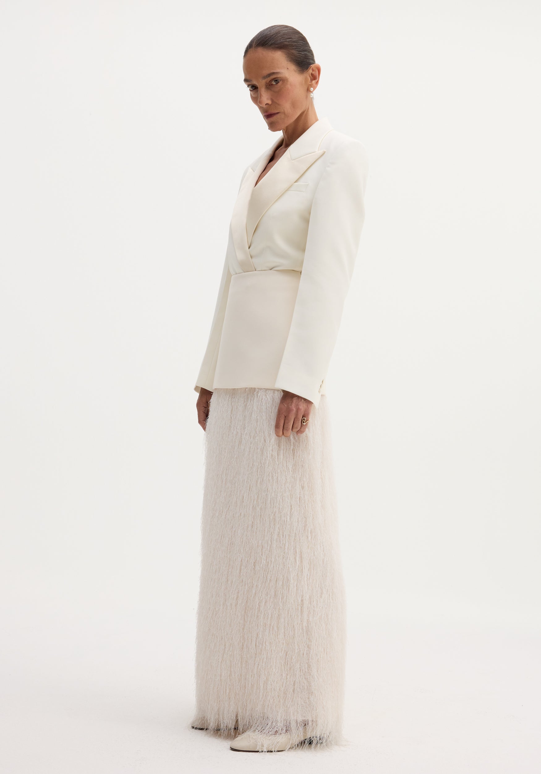 Model wears ivory Fringe skirt with wool upper, full-length fringe detailing, and satin-finish blazer. Accessorized with drop earrings and pointed-toe shoes.