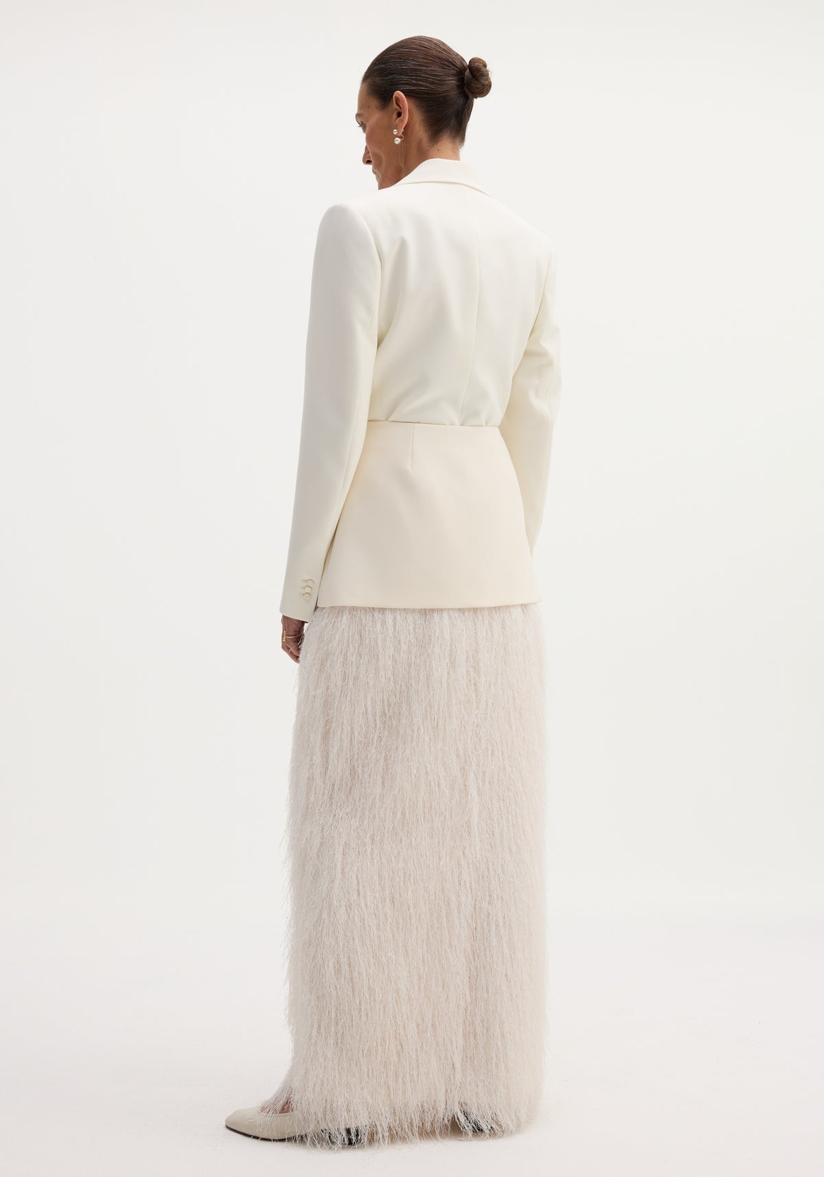 Person wearing an ivory Fringe skirt with full-length eyelash fringe detailing, structured shoulders blazer, pearl drop earrings, and cream heels, viewed from behind.