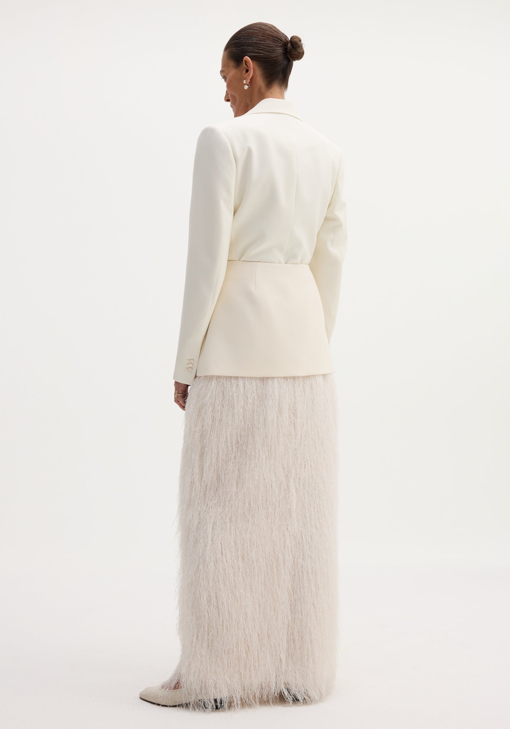 Person wearing an ivory Fringe skirt with full-length eyelash fringe detailing, structured shoulders blazer, pearl drop earrings, and cream heels, viewed from behind.