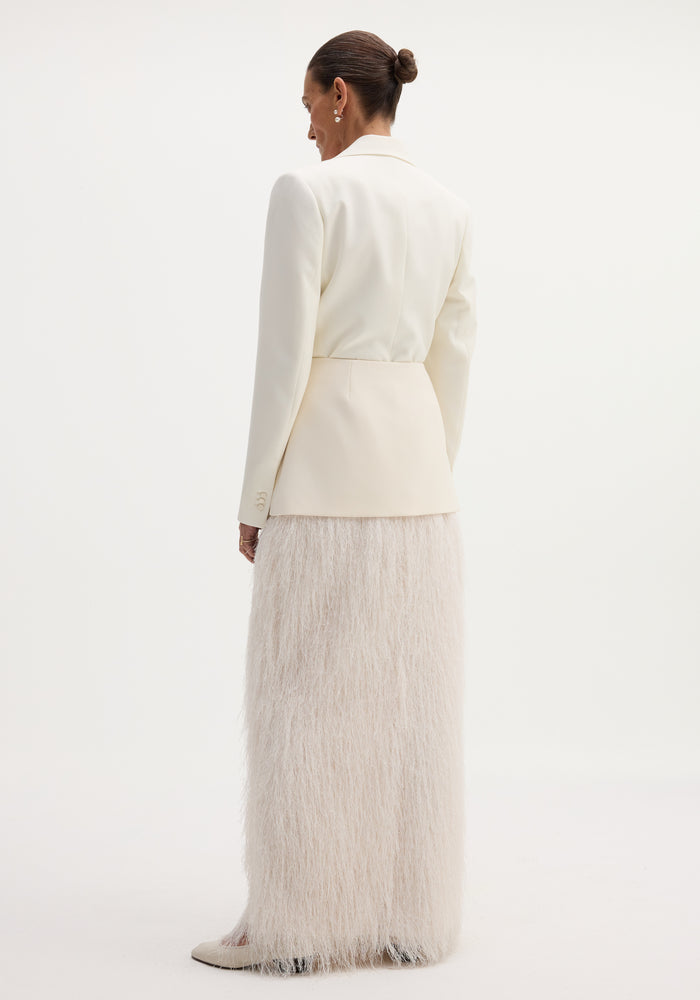 Person wearing an ivory Fringe skirt with full-length eyelash fringe detailing, structured shoulders blazer, pearl drop earrings, and cream heels, viewed from behind.