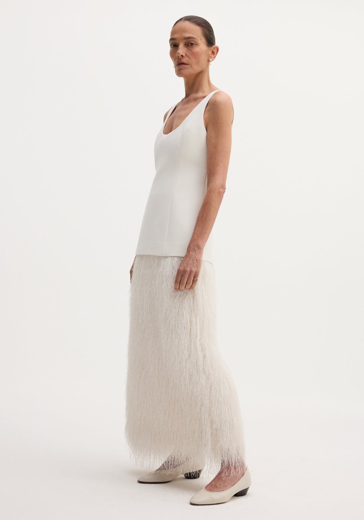 Woman wearing a sleeveless white fringe dress | ivory with a fitted bodice and long fringed skirt, paired with white low-heeled shoes against a plain background.