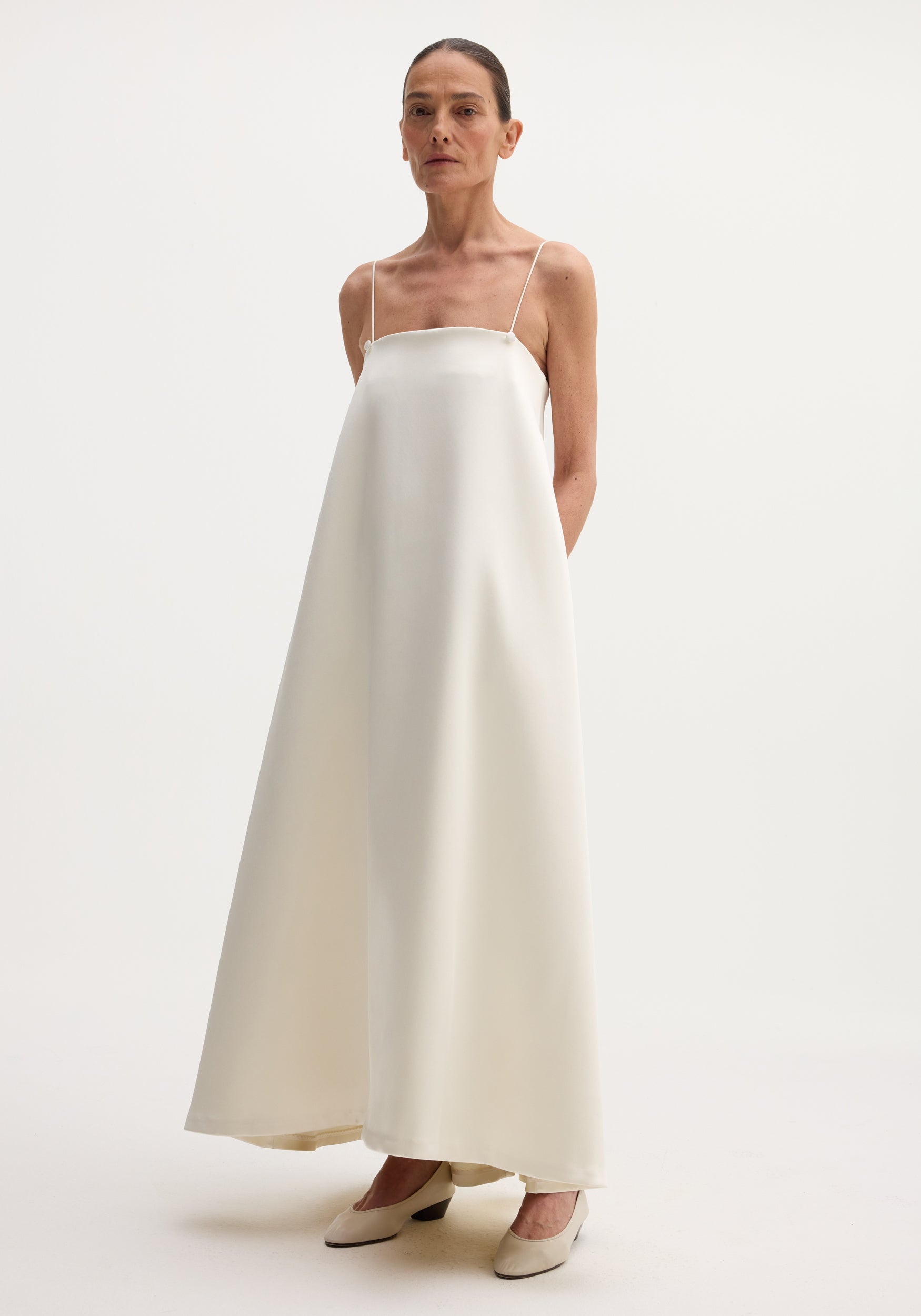 Woman wearing a voluminous satin dress | ivory with thin straps, standing against a plain white background and wearing matching ivory low-heeled shoes.