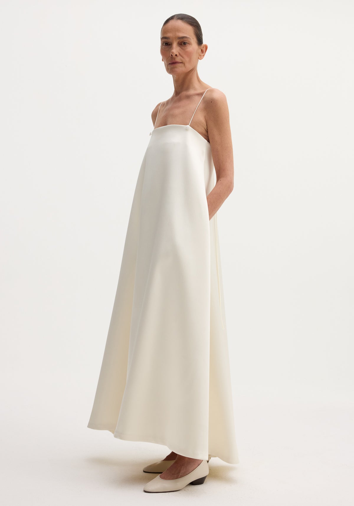 Woman wearing a Voluminous satin dress | ivory with thin straps, paired with matching low-heeled shoes, standing against a plain white background.