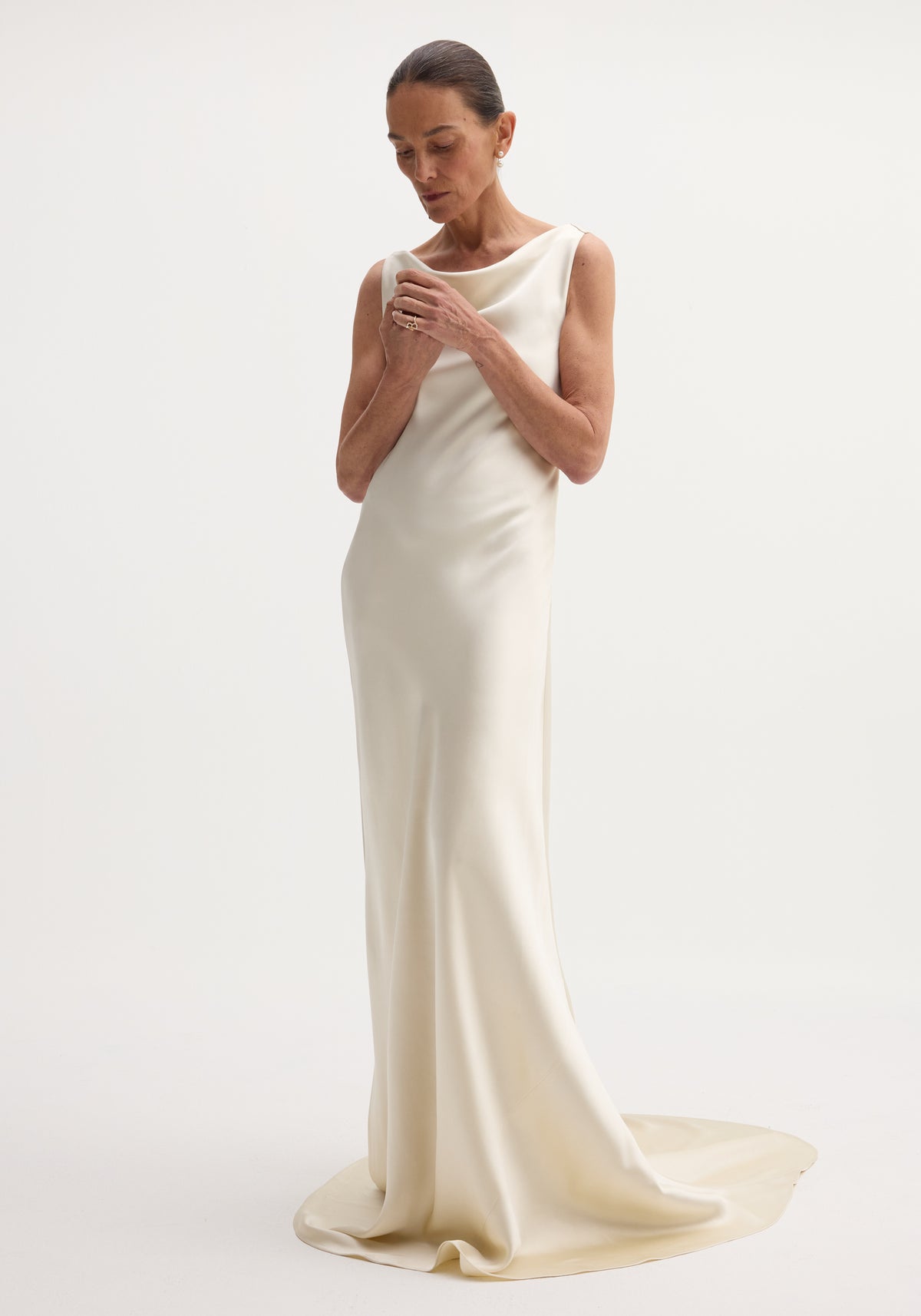 Woman wearing an elegant ivory draped satin dress with train, standing against a plain white background with a minimalist and sophisticated pose, showcasing the smooth fabric and flowing silhouette of the draped satin dress with train | ivory.