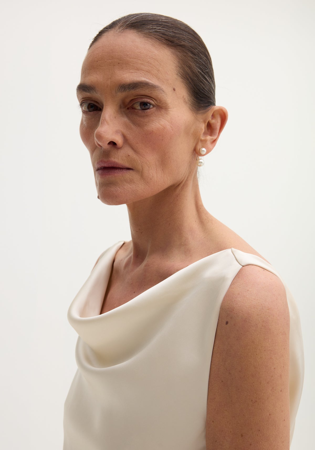 Close-up of a woman wearing a draped satin dress with train | ivory, featuring a soft cowl neckline and sleeveless design, against a plain light background.