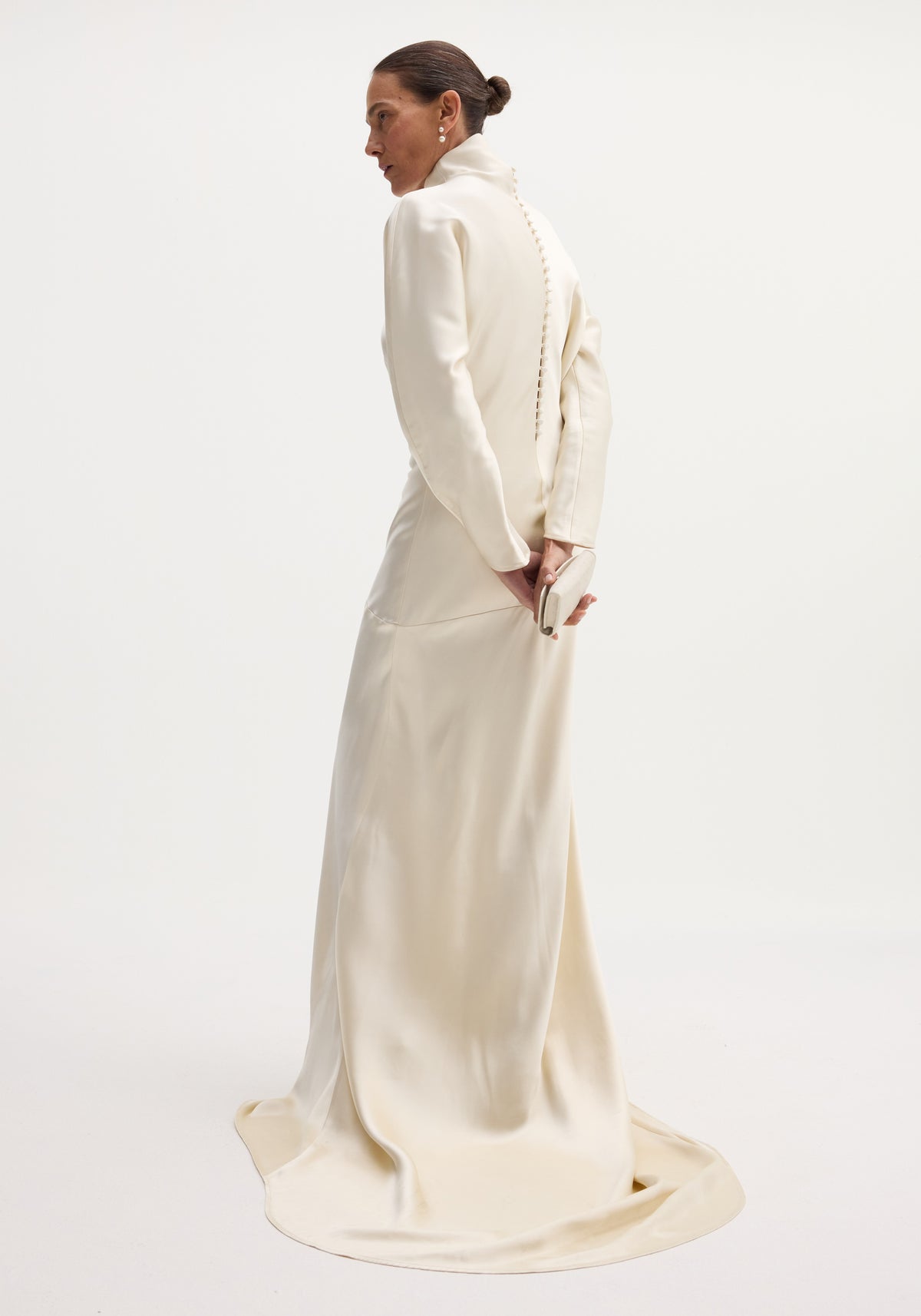 Woman wearing an elegant satin button down dress with train in ivory, featuring long sleeves and a high collar, standing with her hands behind her back against a plain background.