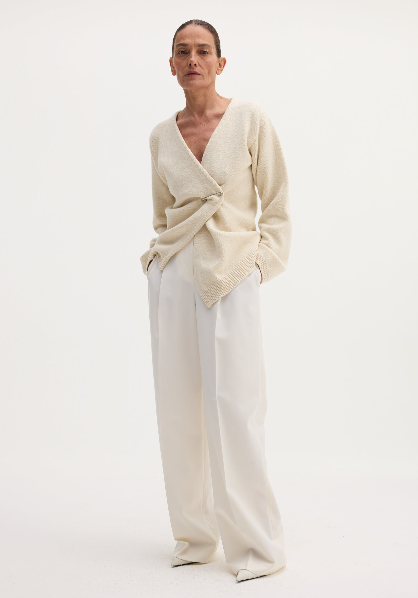 Model wearing Off-shoulder wrap cardigan in cream, featuring a safety-pin closure, paired with high-waisted trousers.
