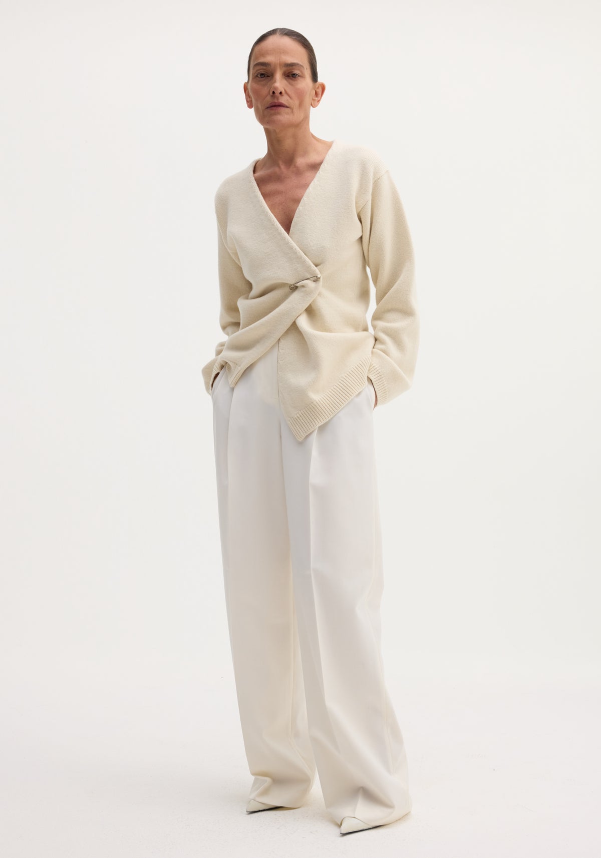 Model wearing Off-shoulder wrap cardigan in cream, featuring a safety-pin closure, paired with high-waisted trousers.