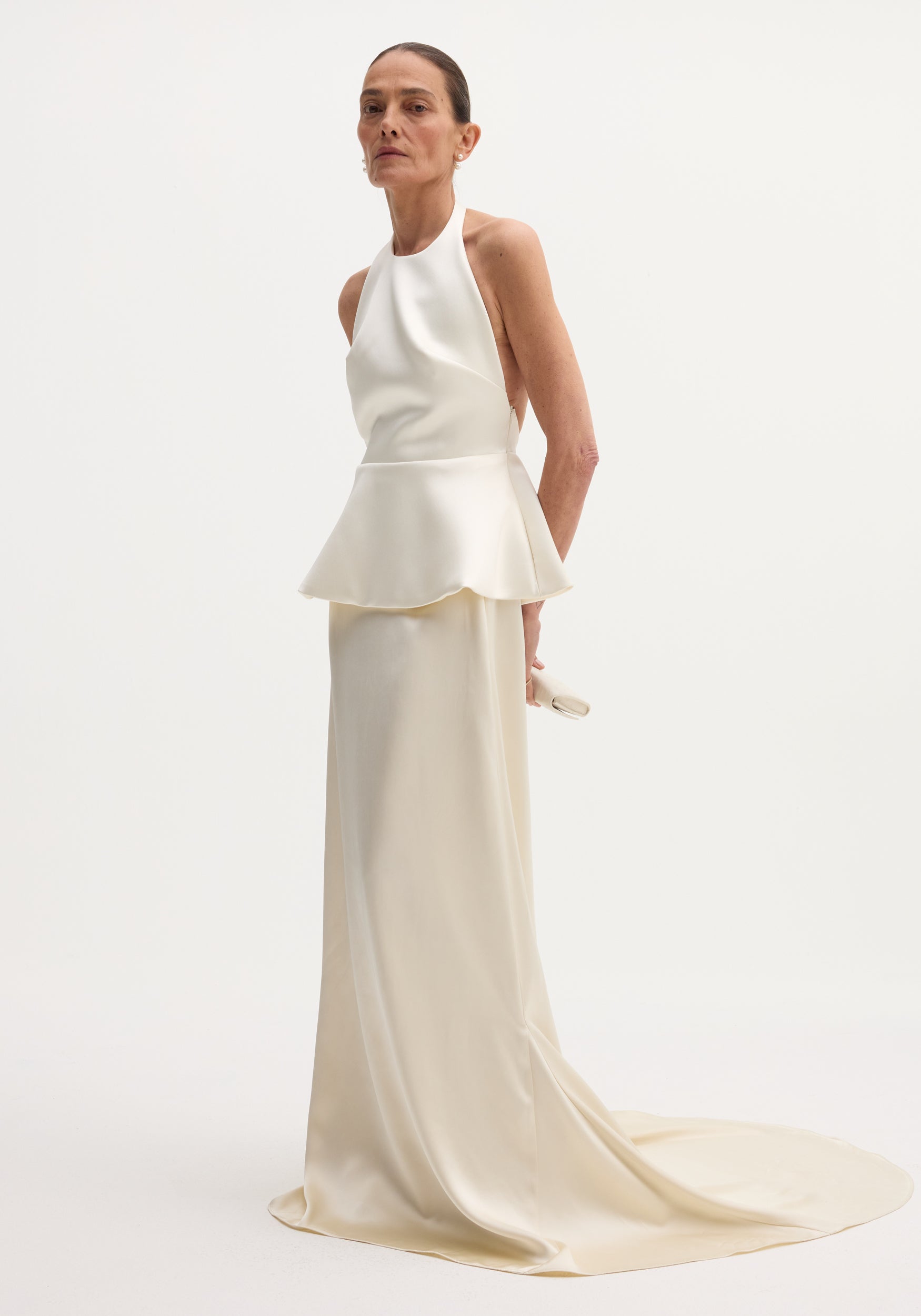 Model wearing sculptural satin halter top with separate silk skirt in ivory, featuring a fitted peplum waist and long flowing skirt with train against a plain background.