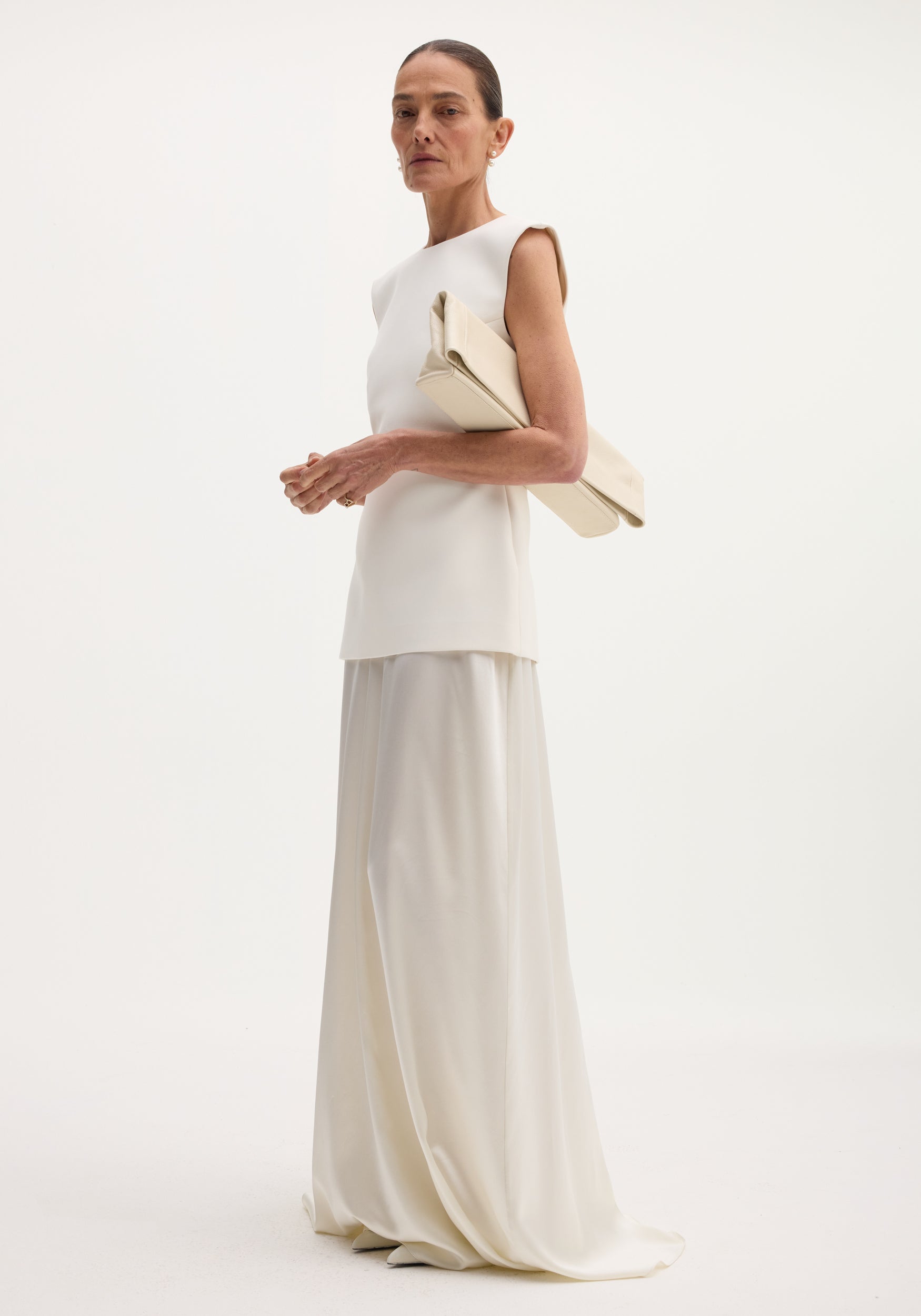 Woman wearing a minimalist ivory sleeveless top paired with a flowing satin silk skirt, holding a matching ivory clutch, showcasing the Classic signature dress with satin silk skirt | ivory.