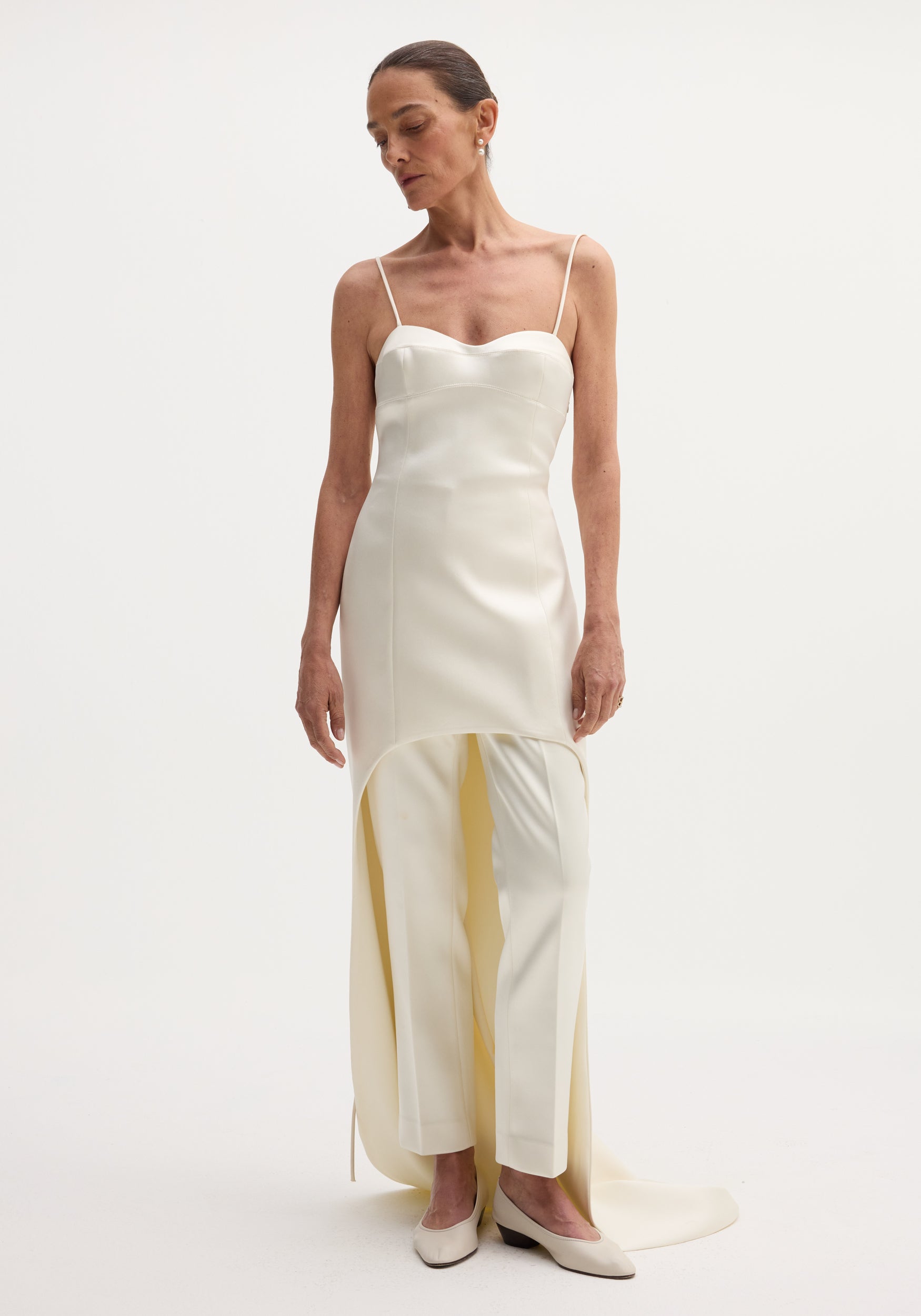 Model wearing the Satin bustier dress with train in ivory, featuring adjustable straps, bustier-style bodice, and asymmetrical drape, paired with straight-leg trousers.