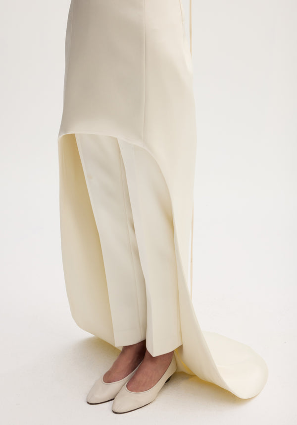 Model wears Satin bustier dress with train, featuring a floor-length, asymmetrical skirt over trousers, and low-heeled shoes in an ivory studio setting.