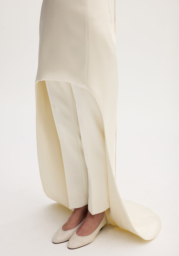 Model wears Satin bustier dress with train, featuring a floor-length, asymmetrical skirt over trousers, and low-heeled shoes in an ivory studio setting.