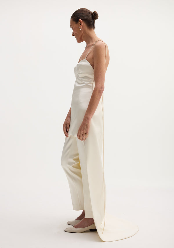 Woman wearing an ivory satin bustier dress with a train, paired with loose trousers, low heels, and drop earrings. Hair in a bun.