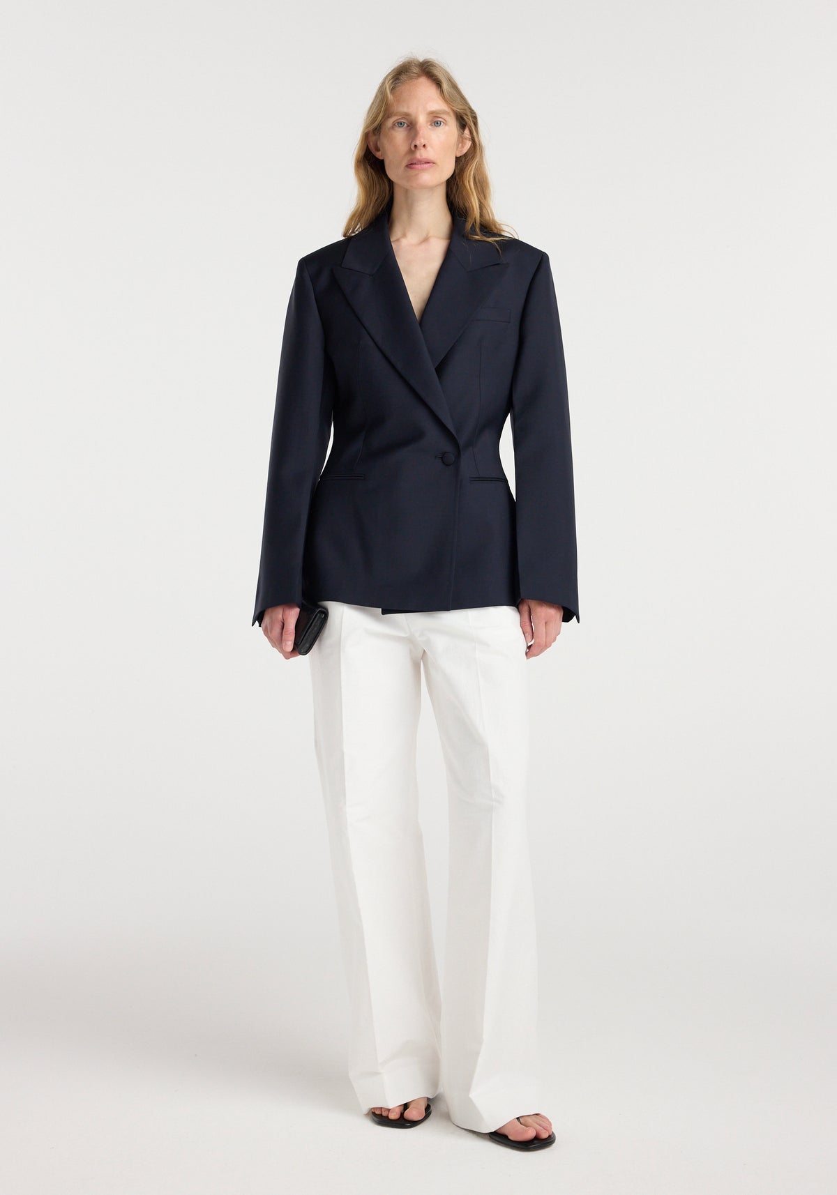Woman wearing an hourglass shaped blazer | navy with wide lapels, paired with white wide-leg trousers and black open-toe shoes against a plain background.