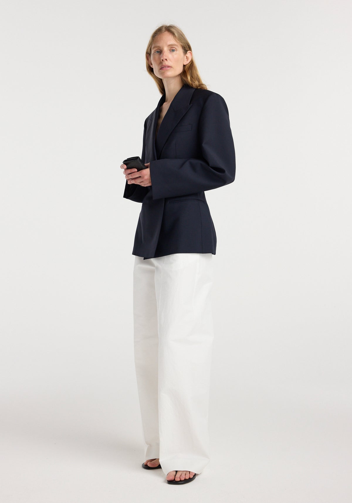 Woman wearing an hourglass shaped blazer | navy with structured shoulders and a tailored fit, paired with wide-leg white pants and black open-toe sandals, holding a phone.