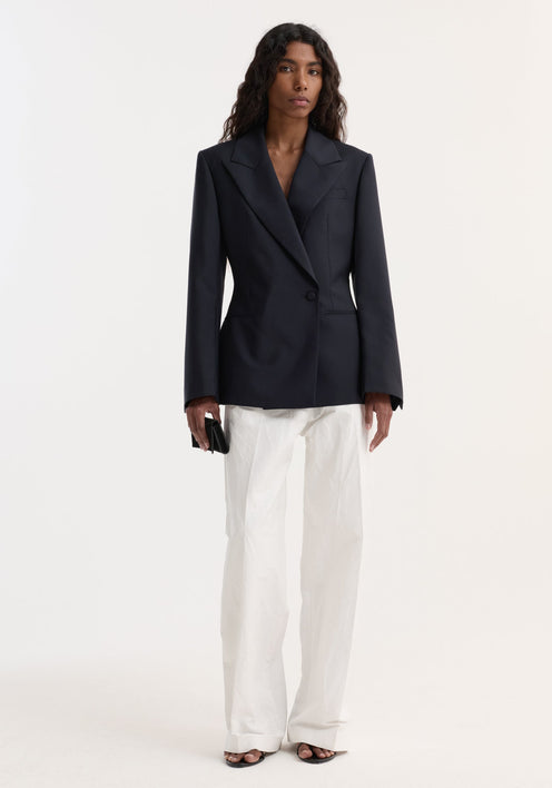 Woman wearing Hourglass shaped blazer with sharp notch lapel, cinched waist, and single button closure, styled with wide-leg trousers, open-toe sandals, and black clutch.