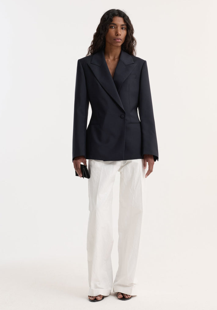 Woman wearing Hourglass shaped blazer with sharp notch lapel, cinched waist, and single button closure, styled with wide-leg trousers, open-toe sandals, and black clutch.