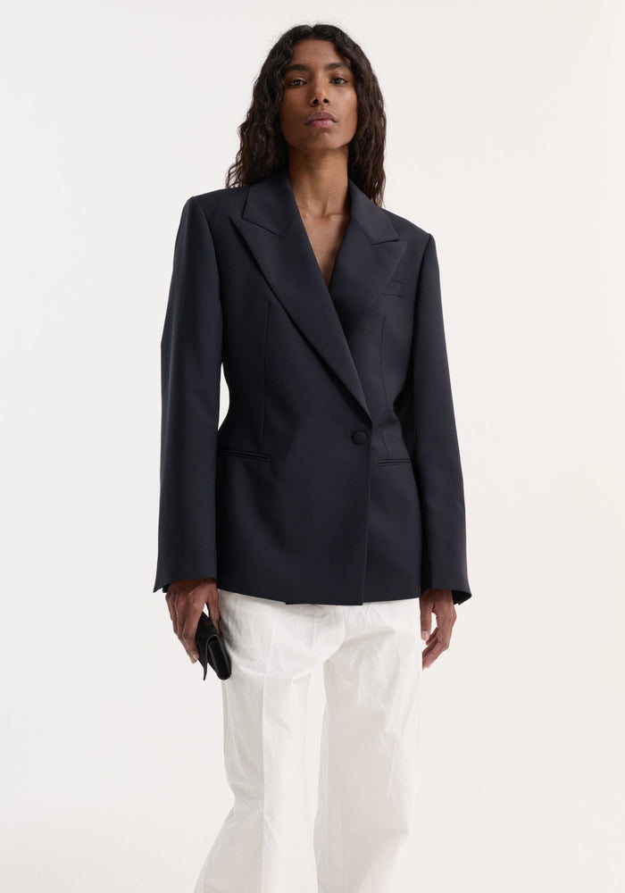 Model in Hourglass shaped blazer with sharp notch lapel, cinched waist, single button closure, paired with loose-cut trousers, holding a black clutch.