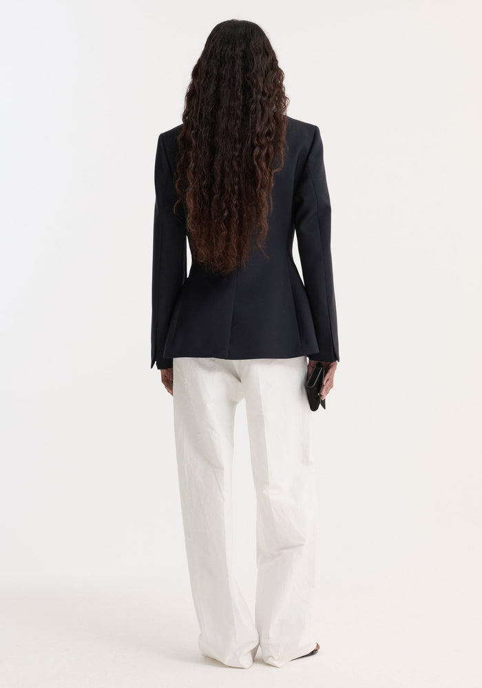 Person wearing an Hourglass shaped blazer with a cinched waist and notch lapel, paired with wide-leg trousers and holding a black clutch.