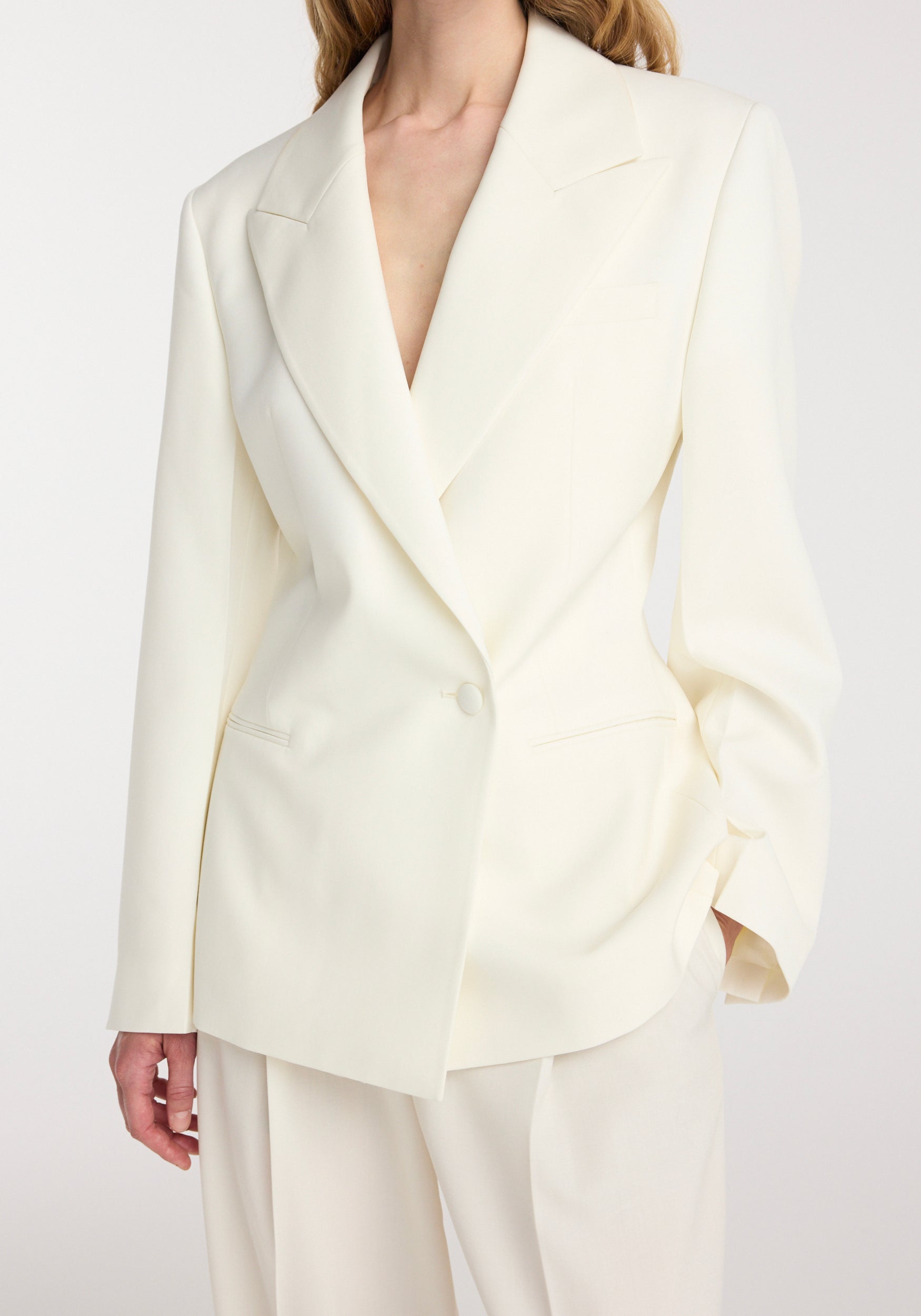 Woman wearing an off white hourglass shaped blazer | off white with a single button closure, peak lapels, and tailored fit, paired with matching loose trousers.