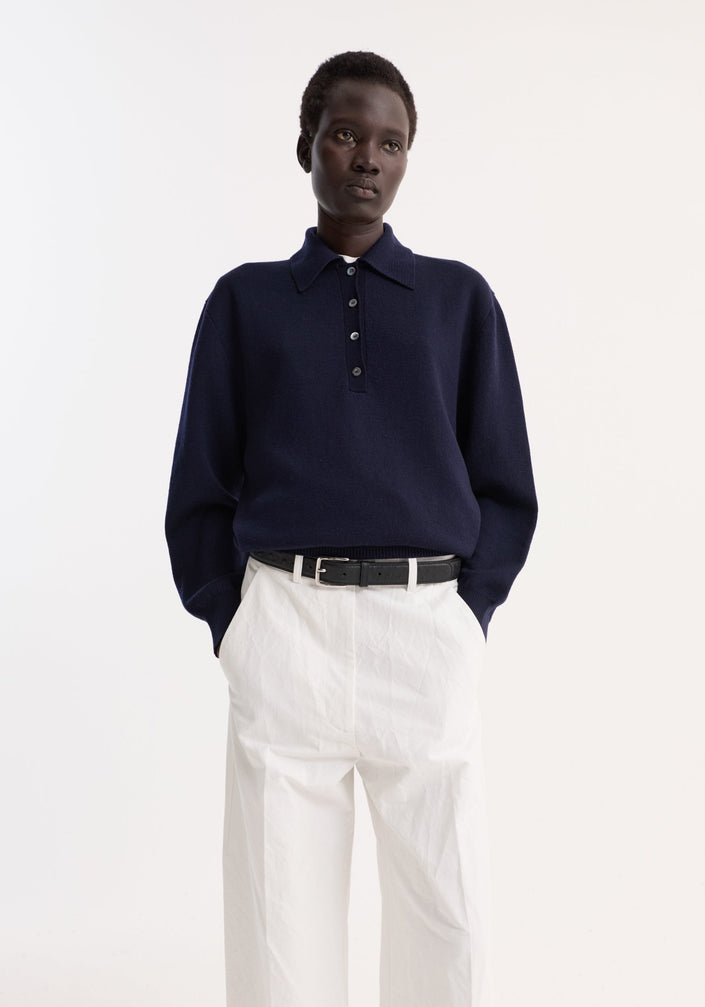 Model in Merino wool polo | navy, showcasing a structured collar and three-button placket, paired with high-waisted white trousers and black sandals.