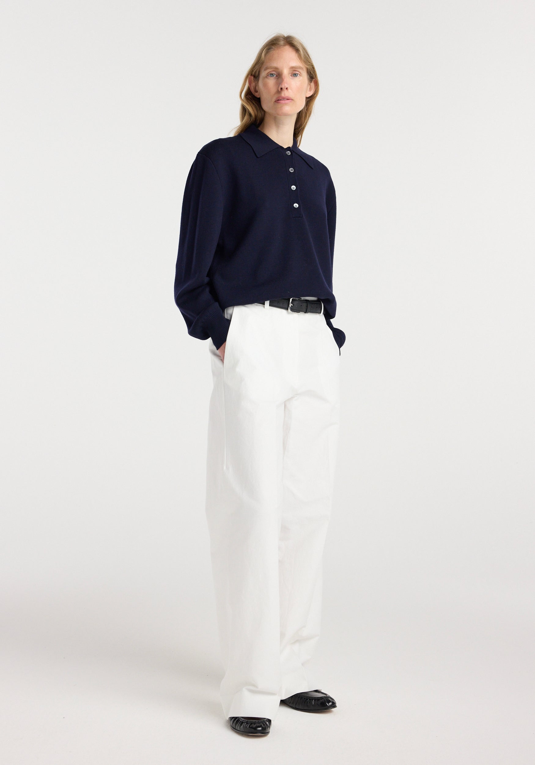 Woman wearing a navy Merino wool polo | navy with long sleeves, paired with white wide-leg pants and black shoes, standing against a plain light background.