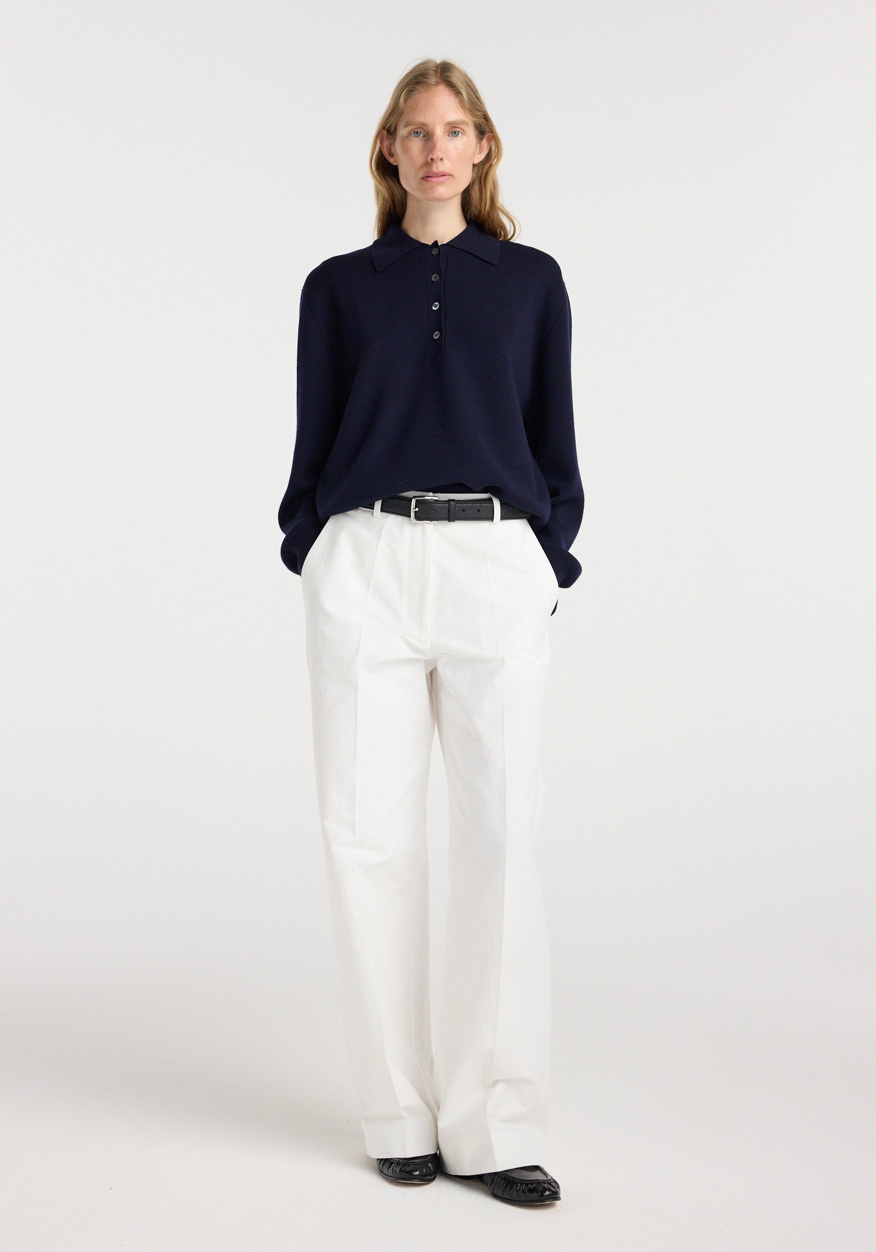 Woman wearing navy Merino wool polo | navy with long sleeves, paired with white high-waisted wide-leg trousers and black belt, standing against plain background.