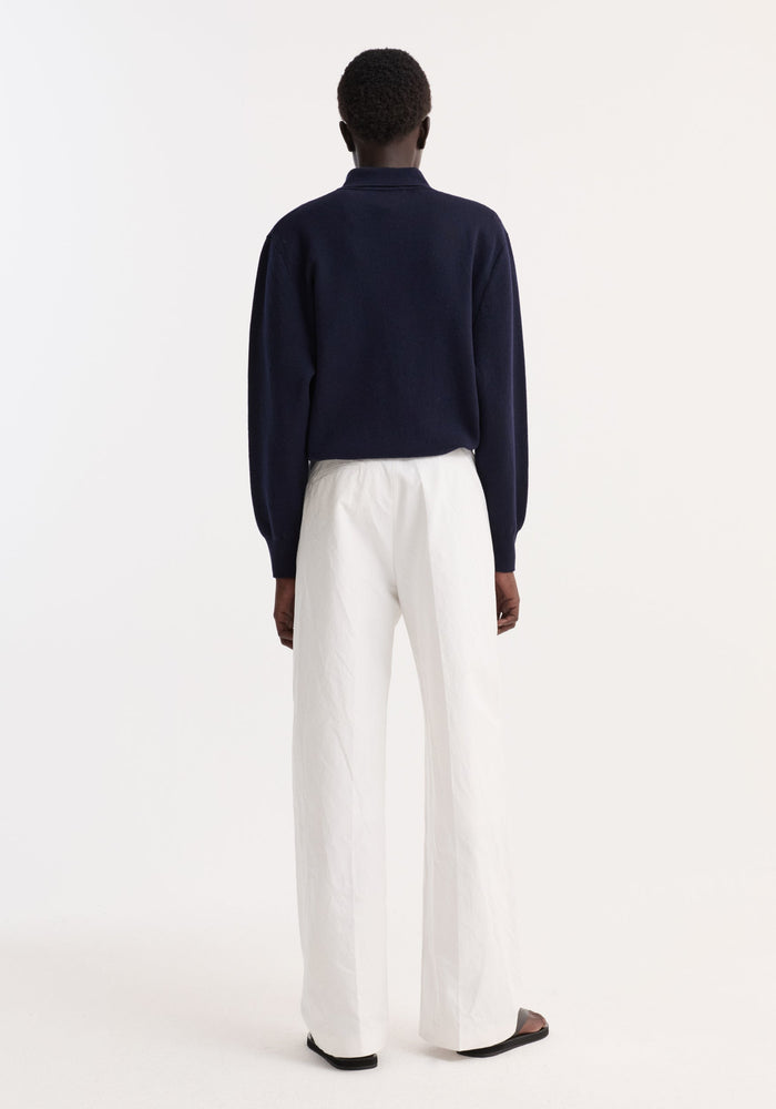 Person in navy Merino wool polo with structured collar and three-button closure, paired with white wide-leg trousers and black sandals, seen from behind.
