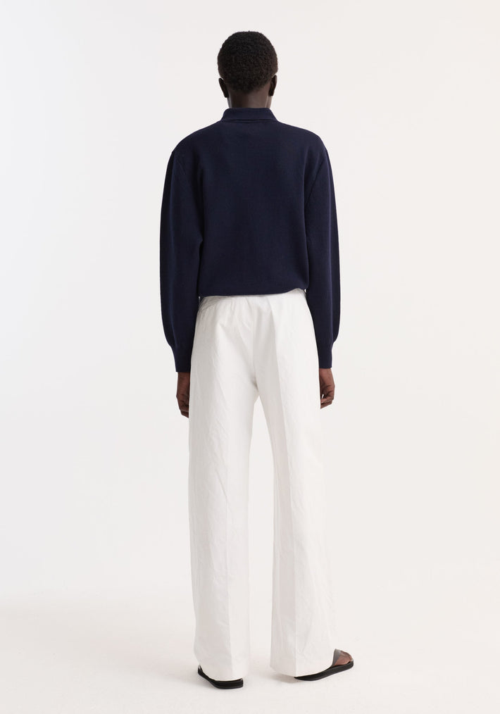 Person in navy Merino wool polo with structured collar and three-button closure, paired with white wide-leg trousers and black sandals, seen from behind.