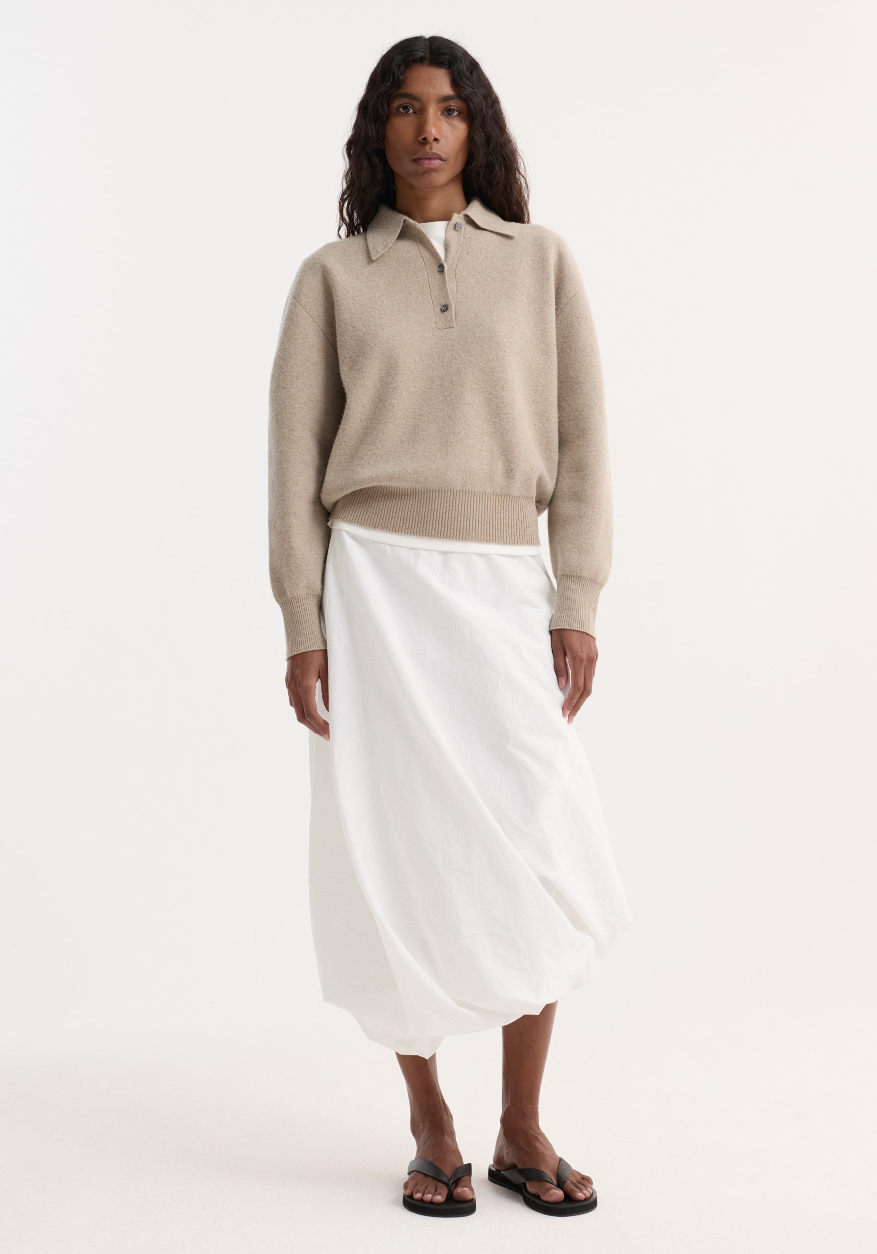 Woman wearing a beige Wool cashmere polo | stone melange with long sleeves and collar, paired with a white midi skirt and black open-toe sandals, standing against a plain background.
