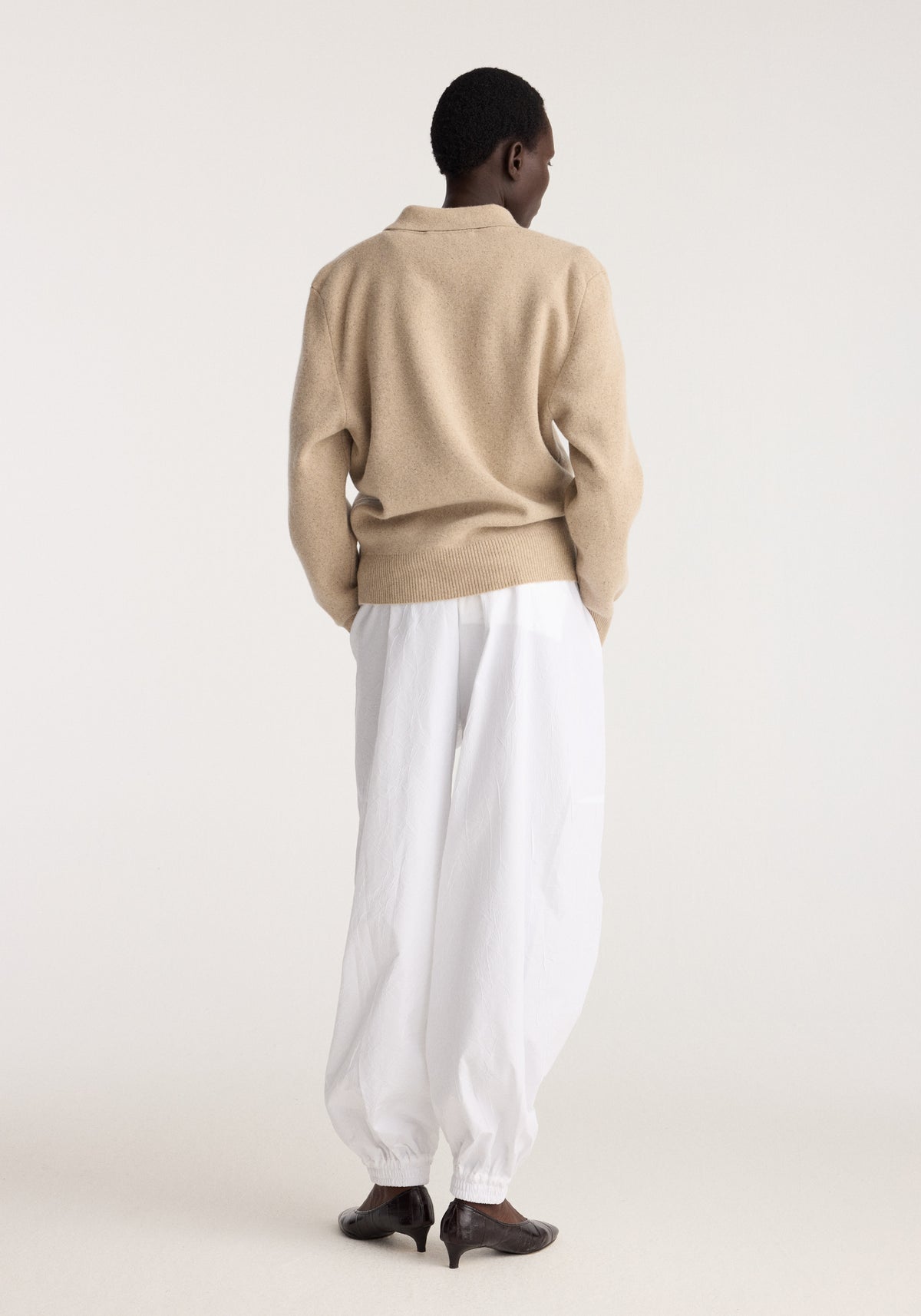 Wool cashmere polo | stone melange seen from the back on a model with white trousers and dark heeled shoes in a neutral studio setting