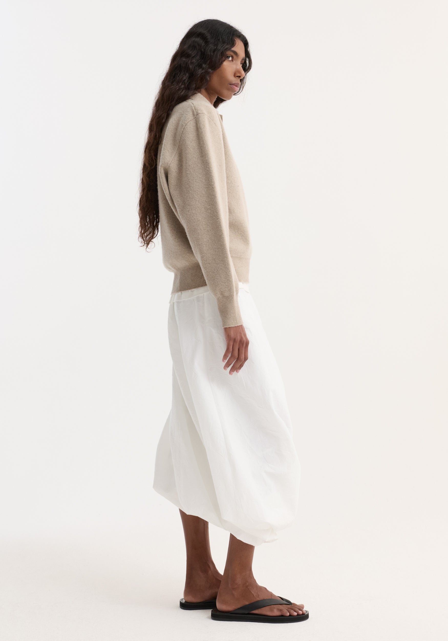 Woman wearing a stone melange Wool cashmere polo paired with a white midi skirt and black sandals, standing in profile against a plain background.